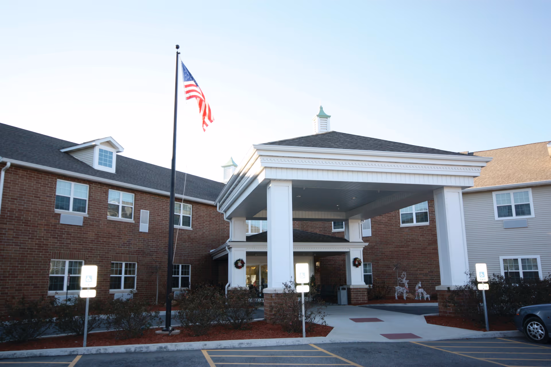 Front entrance of a brick senior living building with a covered porte-cochere and an American flag.