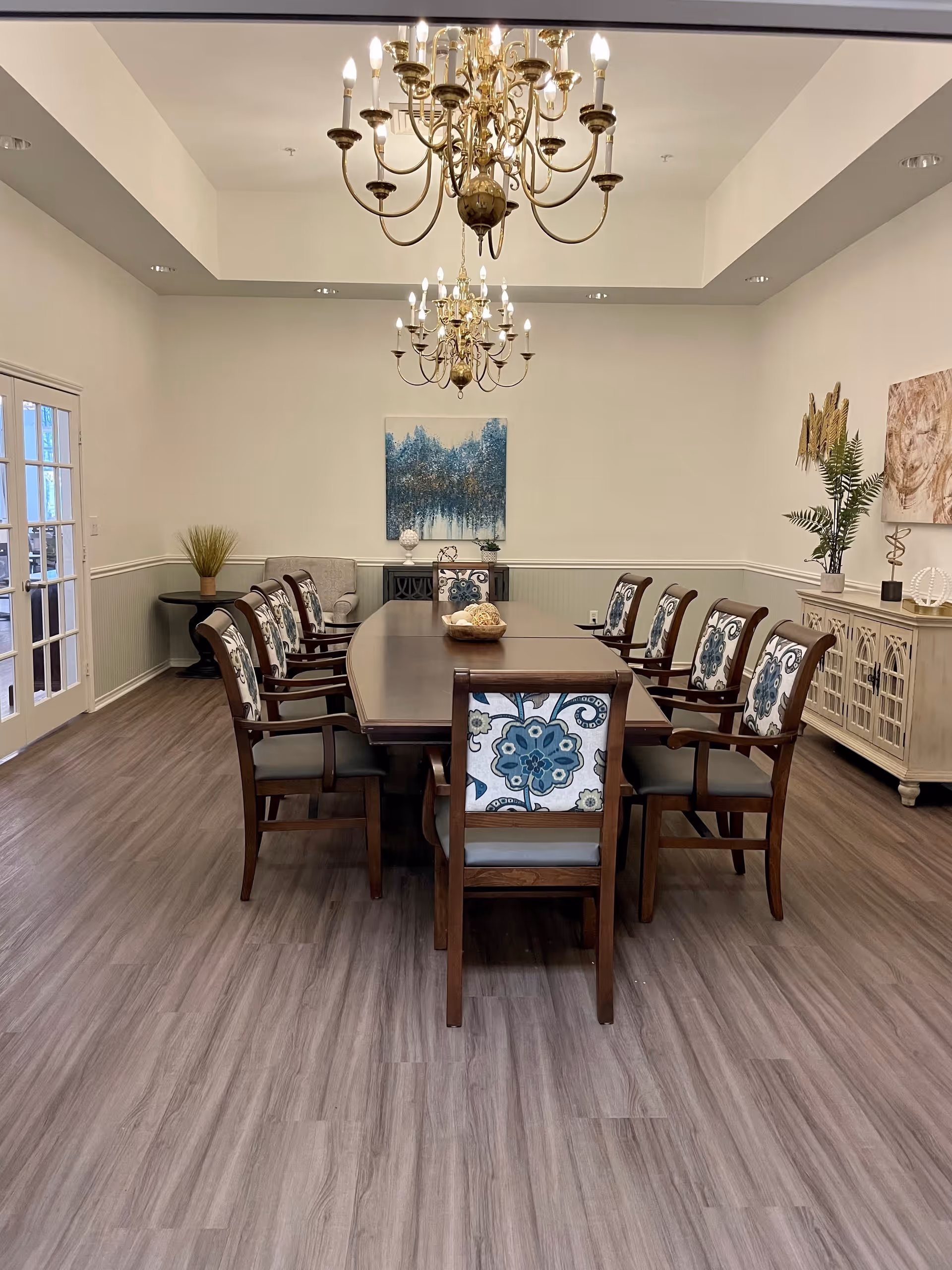 A formal dining room with a long wooden table surrounded by eight wooden chairs with floral patterned cushions. Two ornate chandeliers hang from the ceiling above the table. The room has light-colored walls with wainscoting, a wooden floor, and decorative plants and artwork on the walls and sideboards.