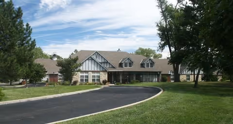 Exterior view of a single-story building with a pitched roof, dormer windows, and a combination of brick and siding. The building is surrounded by green grass, trees, and a curved paved driveway leading to the entrance under a covered porch.