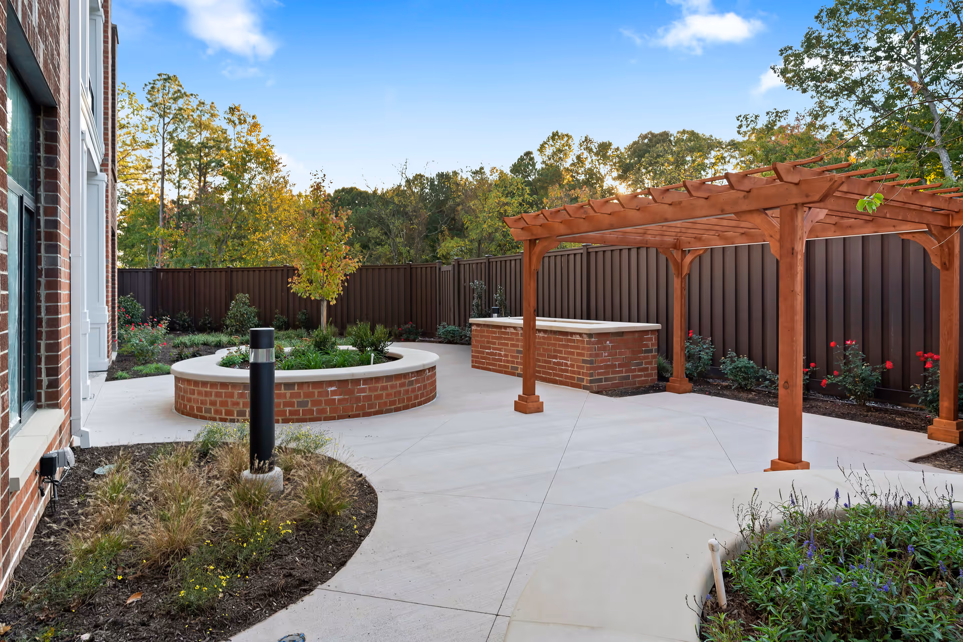 Outdoor patio area at Elancé at Tuckahoe featuring a circular brick planter with greenery, a wooden pergola, a brick counter or bar area, and a concrete walkway surrounded by landscaped plants and trees under a clear blue sky.