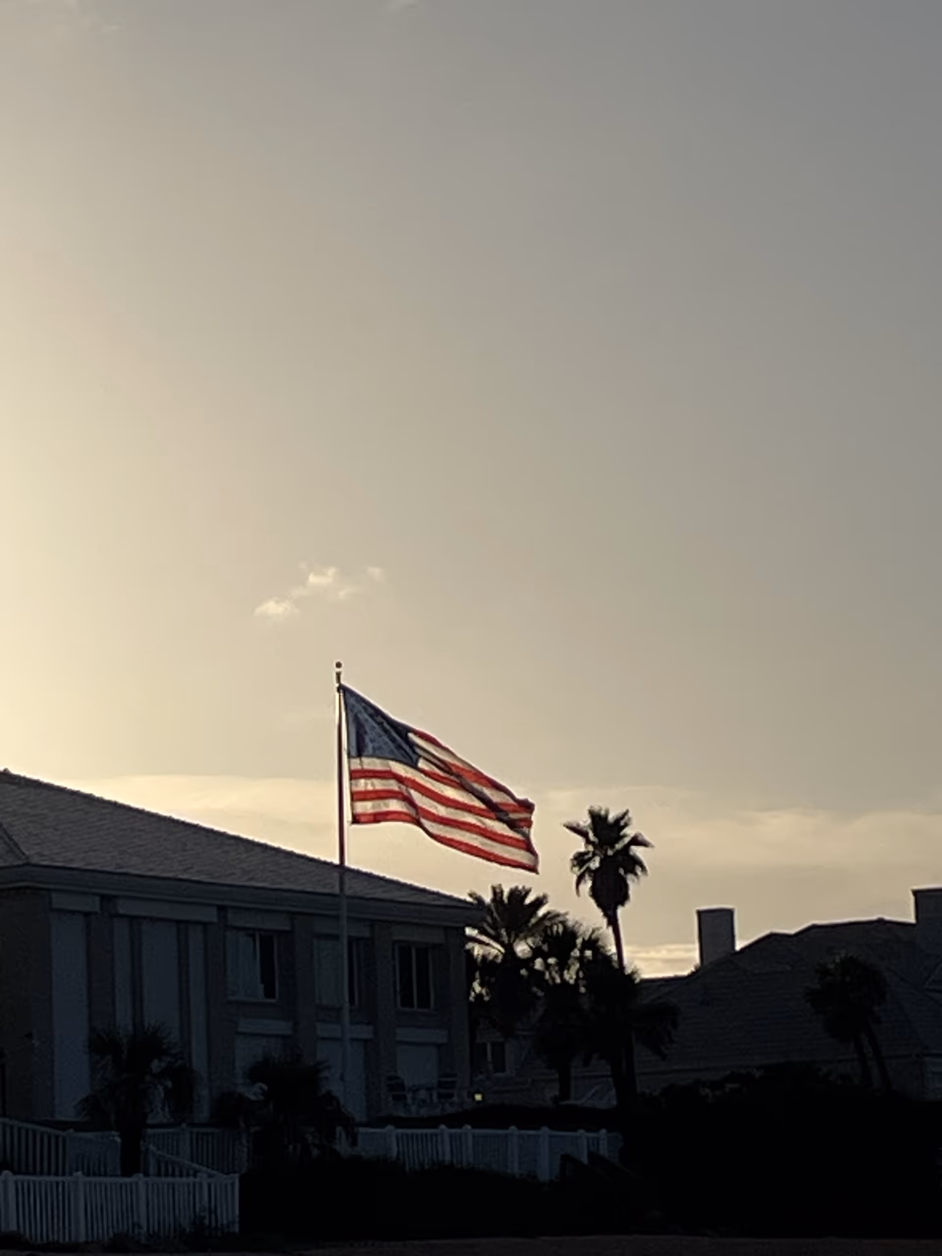 An American flag waves in front of a senior facility with palm trees at sunset.