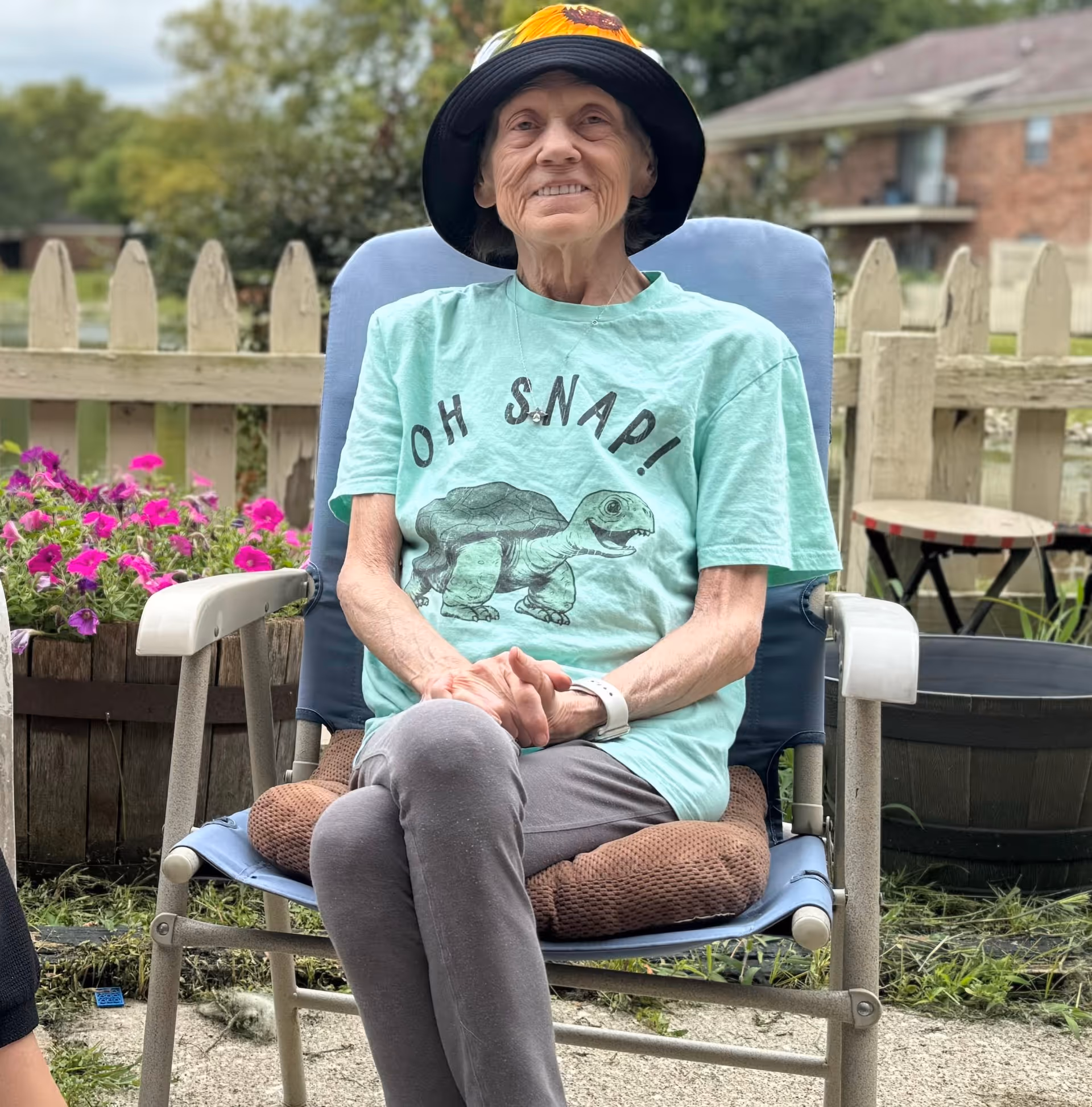 Elderly woman wearing a hat sits in a blue chair outdoors, with flowers and fence behind.