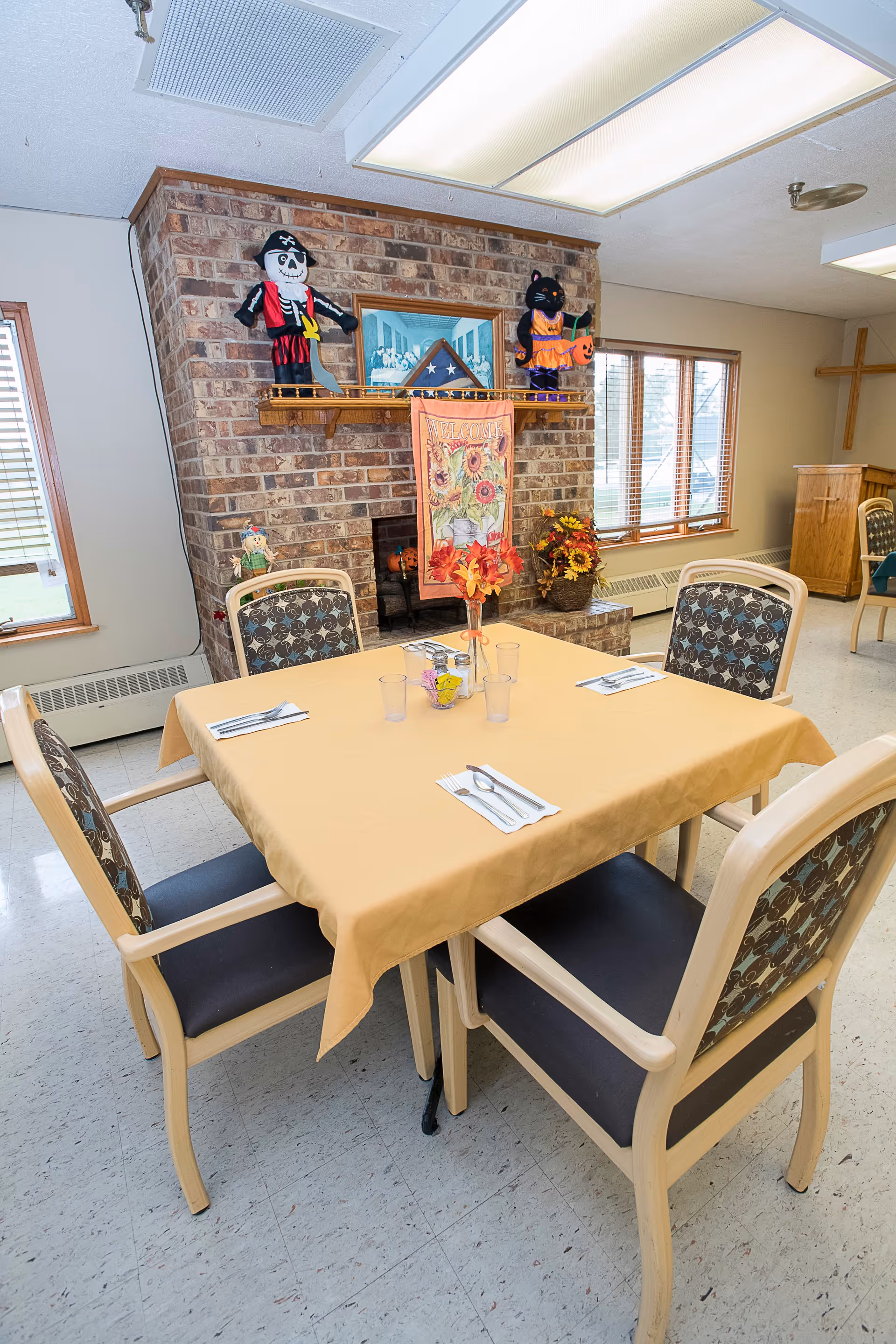 A dining area with a square table covered with a yellow tablecloth set for four people with utensils and cups. The room features a brick fireplace decorated with Halloween-themed items including a pirate skeleton and a black cat holding a pumpkin. There are windows with blinds and a wooden cross mounted on the wall in the background.