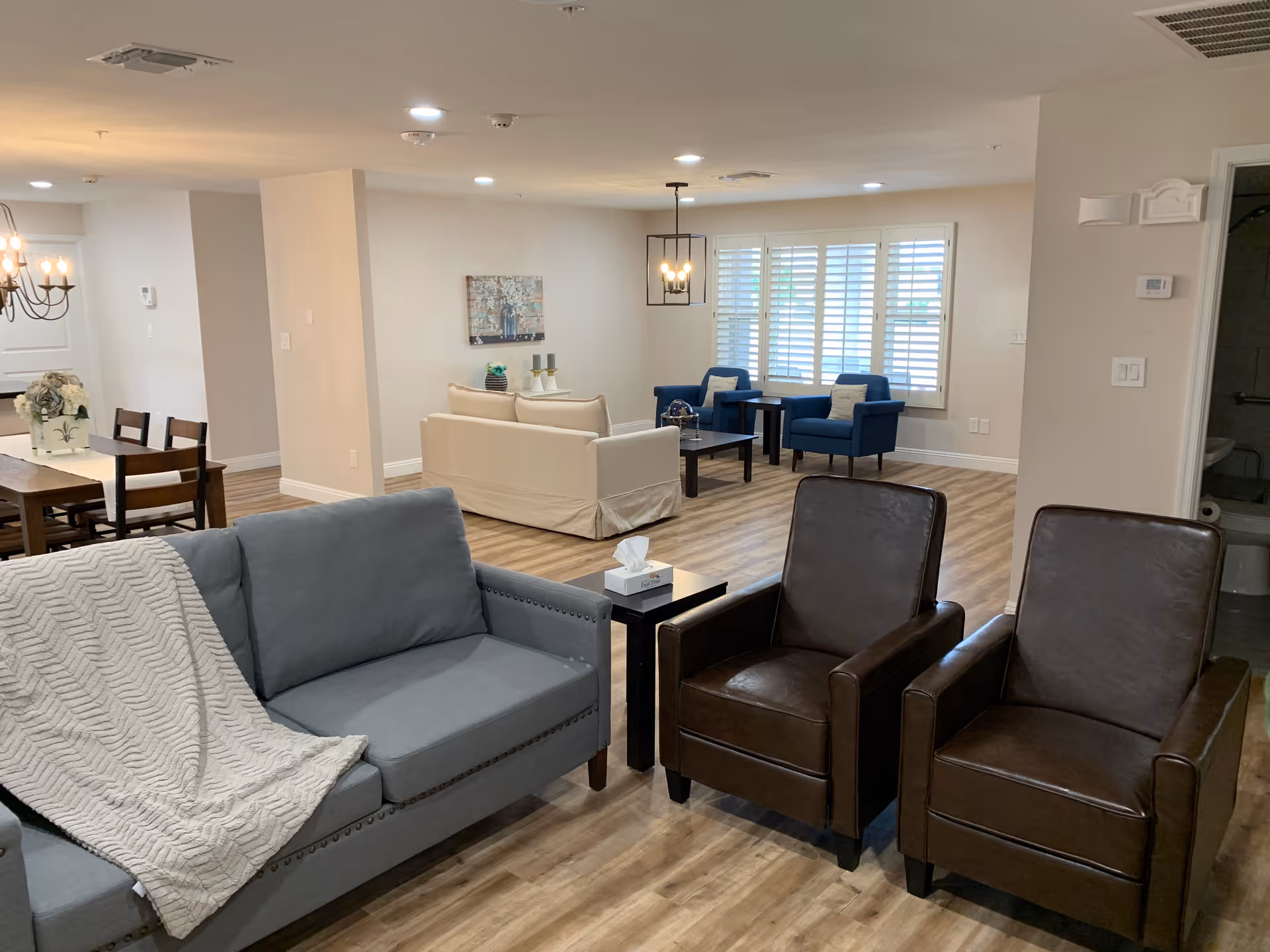 A spacious and well-lit living room area in an assisted living care home featuring a gray sofa with a white knitted throw, two brown leather armchairs, a beige couch, two blue armchairs, wooden coffee tables, and a dining table with chairs in the background. The room has wooden flooring, white walls, and large windows with white shutters allowing natural light to enter.