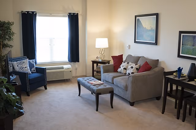 A cozy living room with a gray sofa adorned with red and patterned pillows, a small rectangular ottoman in front, a blue armchair near a window with dark curtains, a side table with a lamp, framed artwork on the walls, and a small dining table with two chairs.