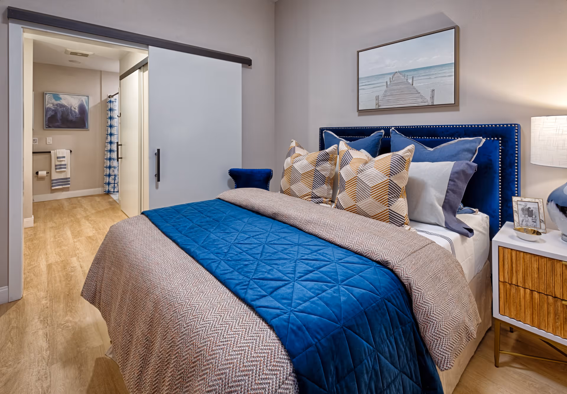 A cozy bedroom with a bed featuring a blue quilt and multiple patterned pillows. To the right of the bed is a nightstand with a lamp and framed photos. A framed picture of a pier over water hangs above the bed. In the background, an open sliding door reveals a bathroom with a shower curtain and towel rack.