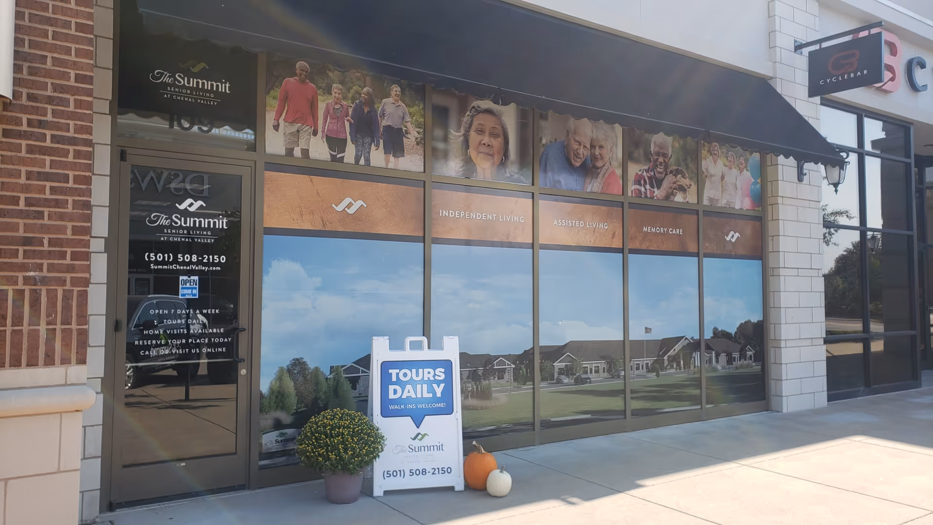 Exterior view of The Summit Senior Living at Chenal Valley facility storefront with large windows displaying images of elderly people and text indicating Independent Living, Assisted Living, and Memory Care. A sandwich board sign outside advertises daily tours with contact information.