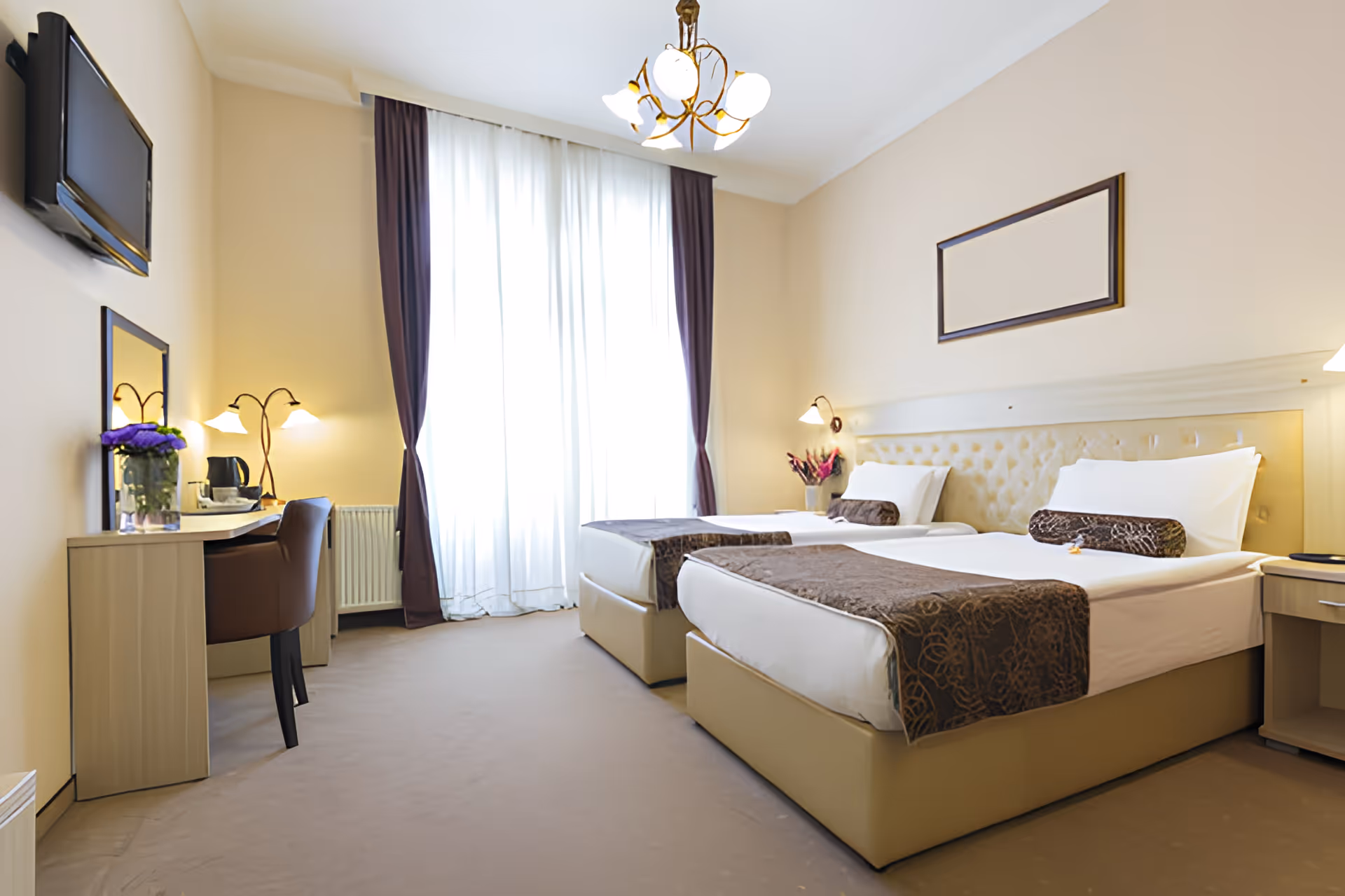Bright twin-bedded bedroom with two single beds, a desk and chair, wall-mounted TV, and large curtained window.