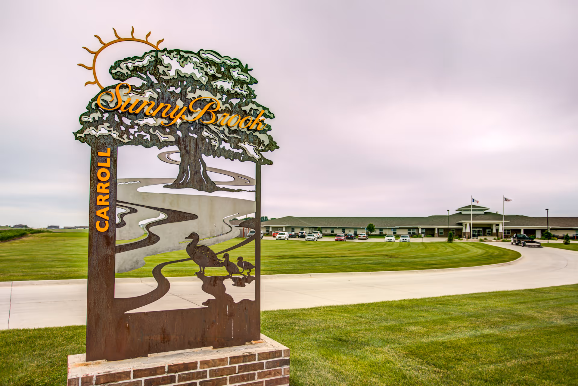 Outdoor view of Sunny Brook Carroll senior living facility with a decorative metal sign featuring a tree, sun, and ducks in the foreground and the building with a parking lot in the background under a cloudy sky.