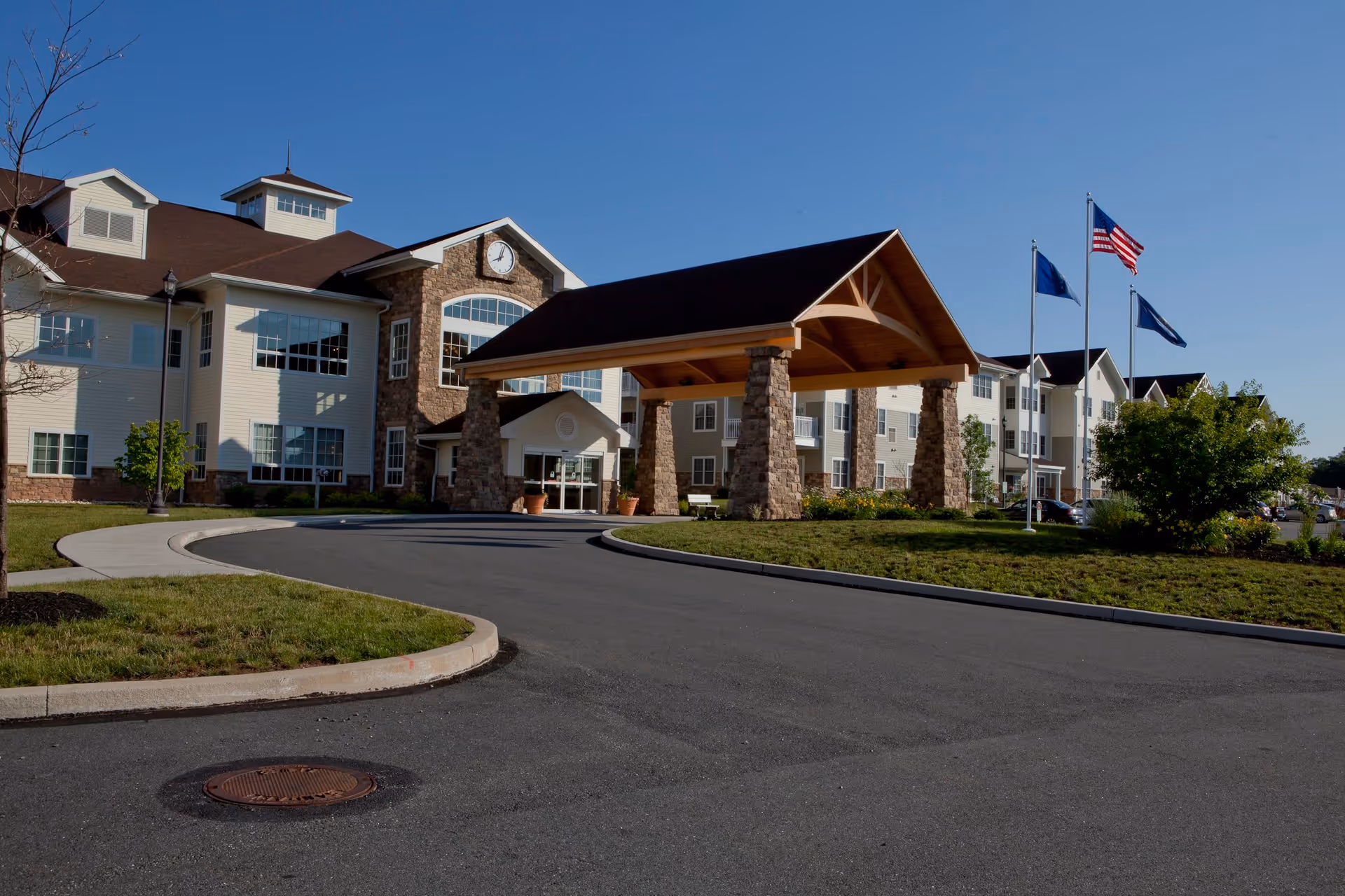 Front exterior view of The Heritage of Green Hills facility showing a large covered entrance supported by stone pillars, a clock above the entrance, multiple windows, a curved driveway, and three flagpoles with flags against a clear blue sky.