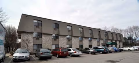 Two-story beige senior living building with parked cars in front and leafless trees nearby.