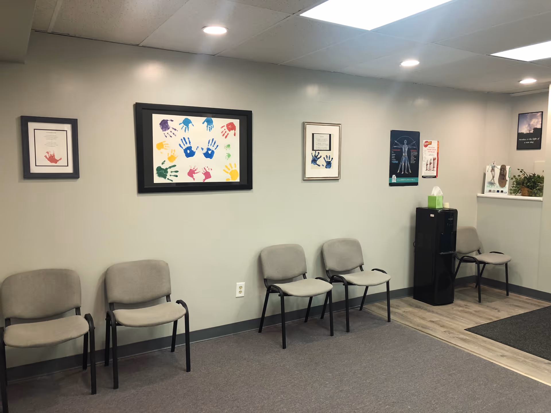 Interior waiting area with several gray chairs and framed posters along the wall.