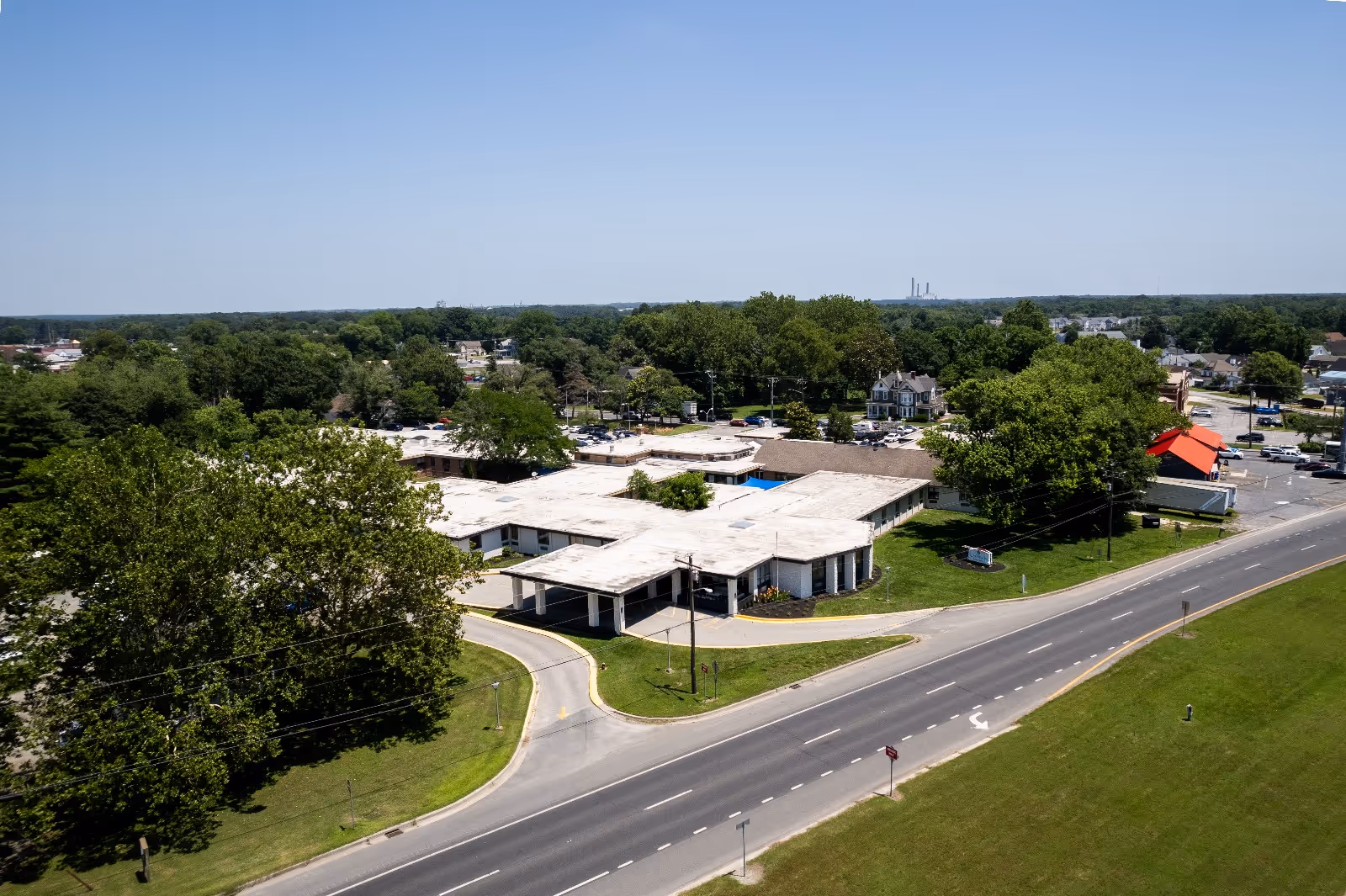 Aerial view of a single-story post-acute facility with white flat roofs, a circular driveway, surrounding trees and adjacent road.