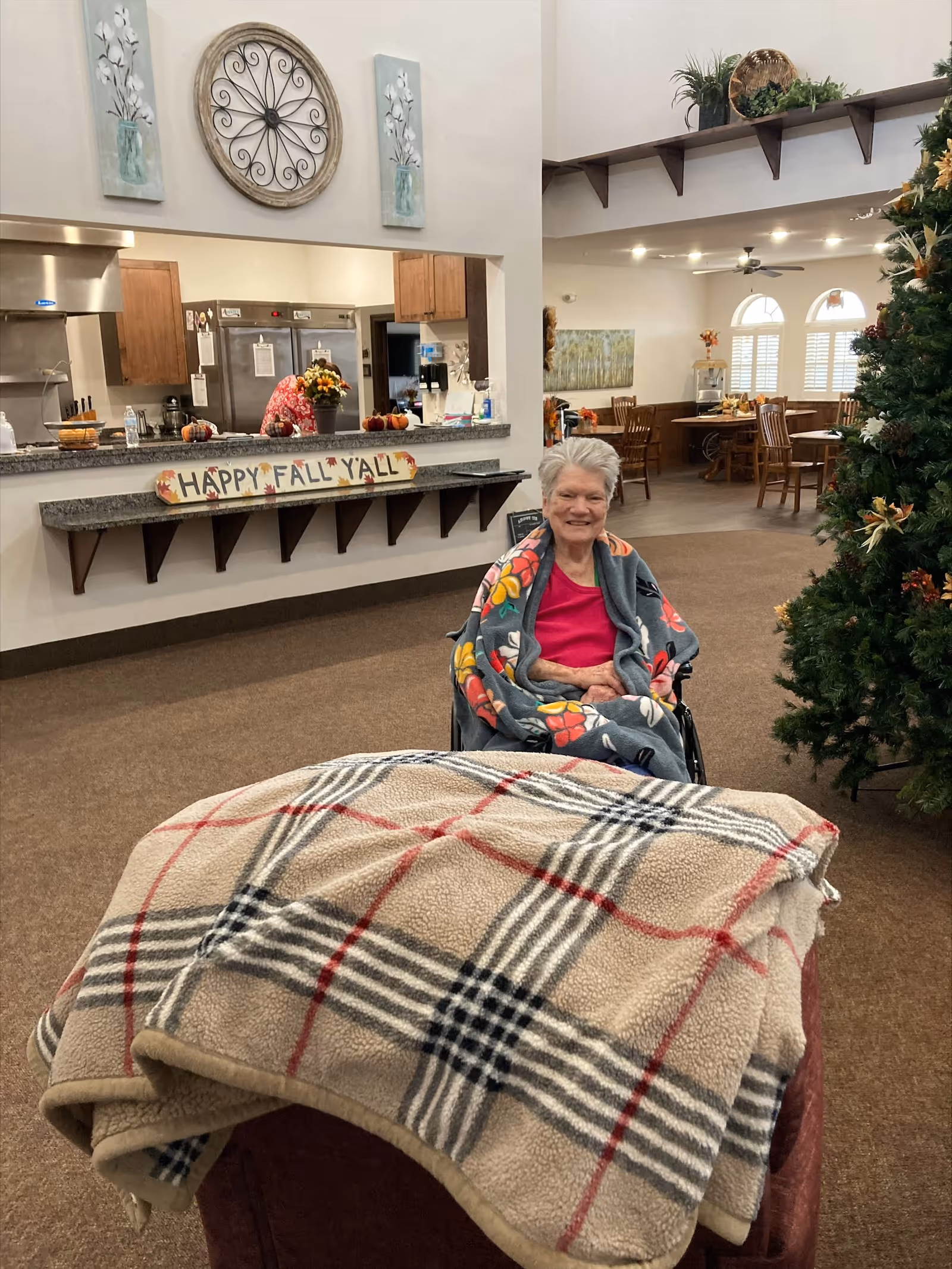 An elderly woman sitting in a wheelchair wrapped in a floral blanket, smiling in a cozy common area of a senior living facility. Behind her is a kitchen counter with a 'Happy Fall Y'all' sign, a person working in the kitchen, and a decorated Christmas tree to the right. The room has warm lighting, wooden furniture, and fall-themed decorations.