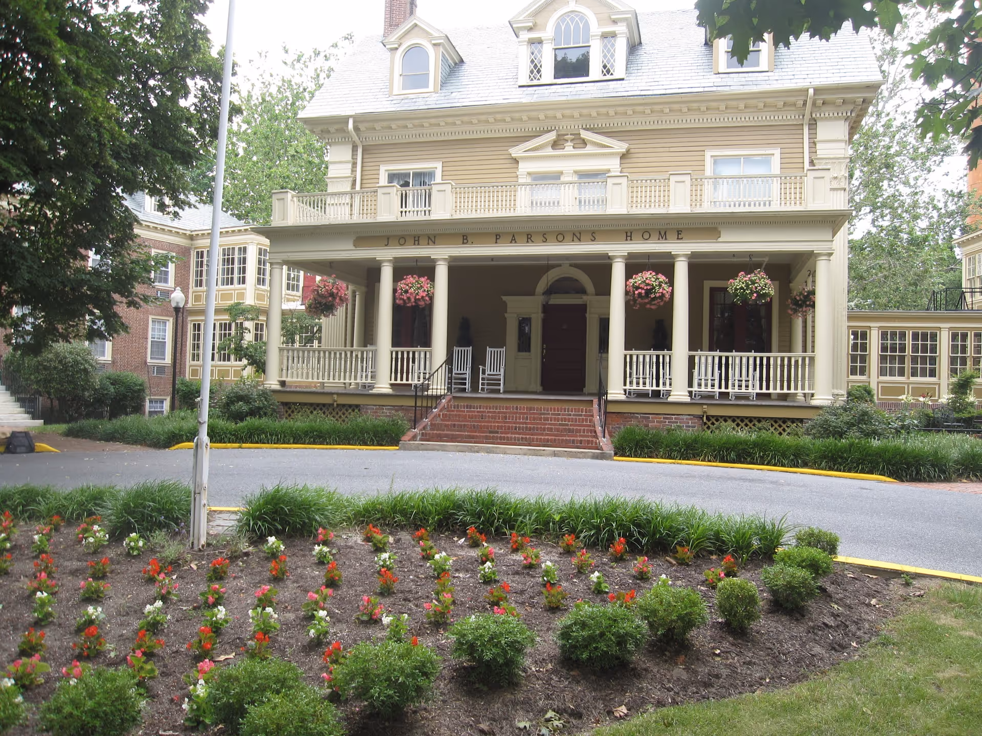 Front exterior view of the John B. Parsons Home, a large historic-style building with a porch featuring columns, hanging flower baskets, and rocking chairs. In front of the building is a circular driveway with a landscaped flower bed containing red and white flowers and green shrubs.