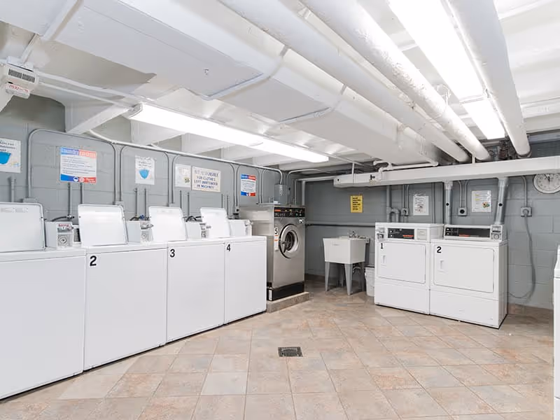 Laundry room with five white washing machines and two white dryers, a utility sink, and various signs on the gray cinder block walls. The ceiling has exposed white pipes and fluorescent lighting.