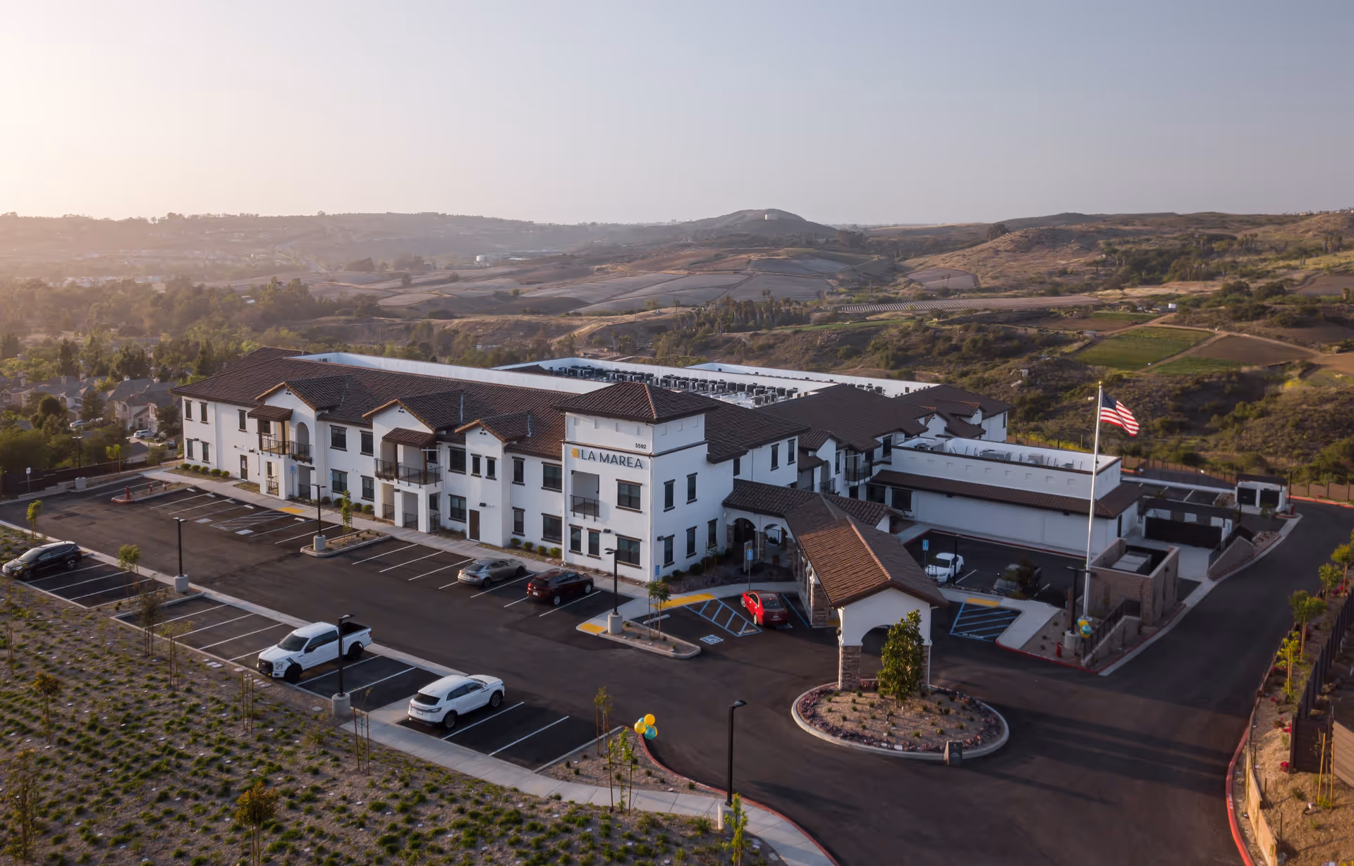 Aerial view of La Marea Senior Living facility showing a large two-story building with white walls and brown roof tiles, surrounded by a parking lot with several cars. The facility is situated in a hilly landscape with greenery and open land in the background under a clear sky.