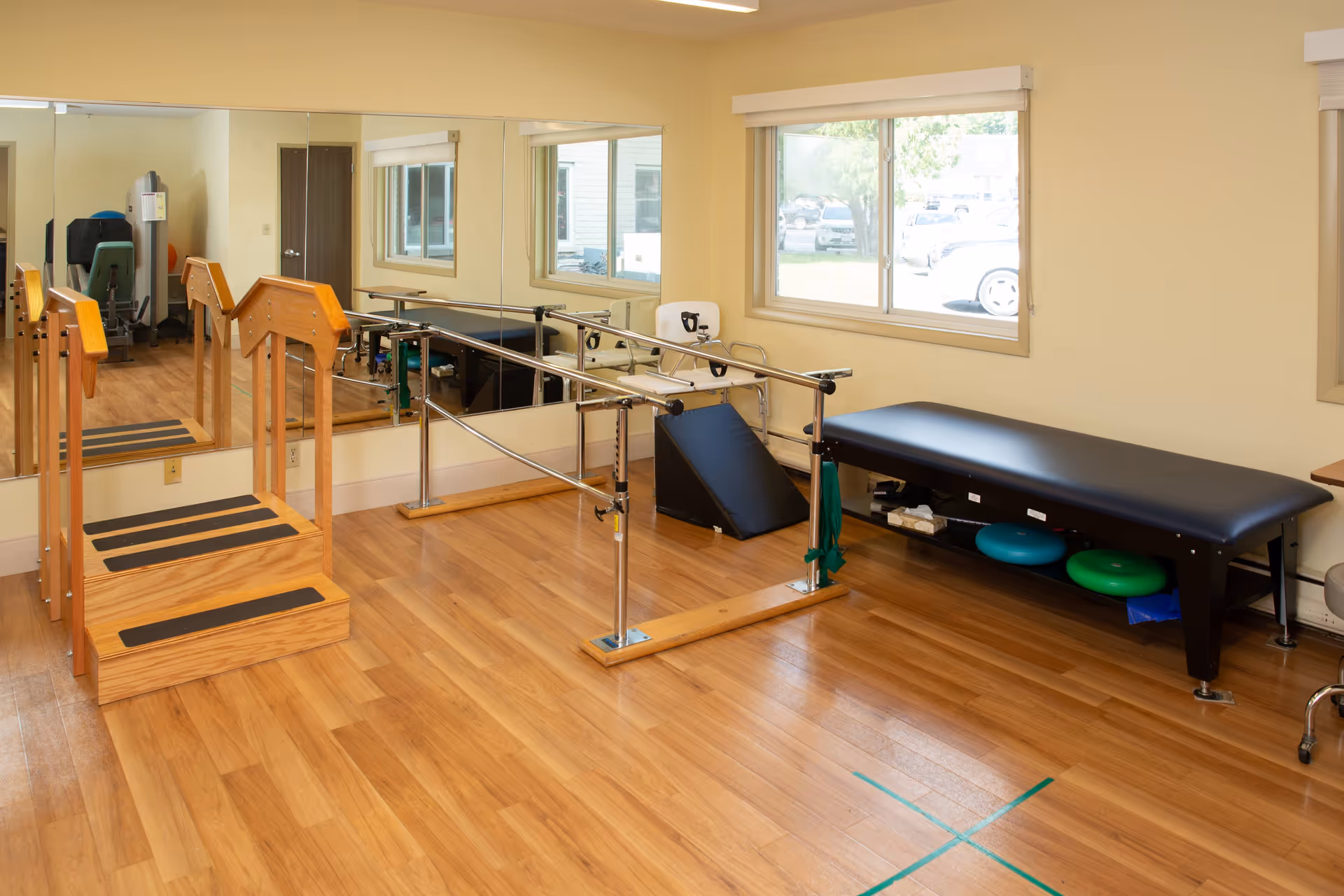 Interior rehab room with parallel bars, a padded treatment table, mirrors, and wooden floors.