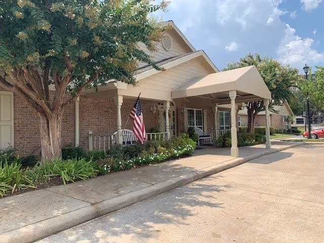 Exterior view of a single-story brick building with a covered entrance supported by columns. There are trees and shrubs along the walkway, an American flag displayed near the entrance, and a clear blue sky with some clouds.