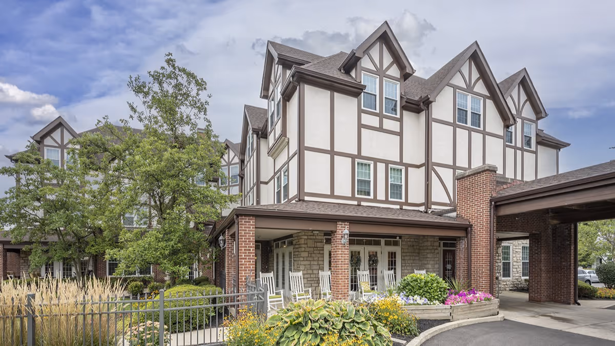 Exterior view of Brookdale Oakwood senior living facility showing a multi-story building with Tudor-style architecture, a covered entrance with white rocking chairs, landscaped greenery, and a partly cloudy sky.