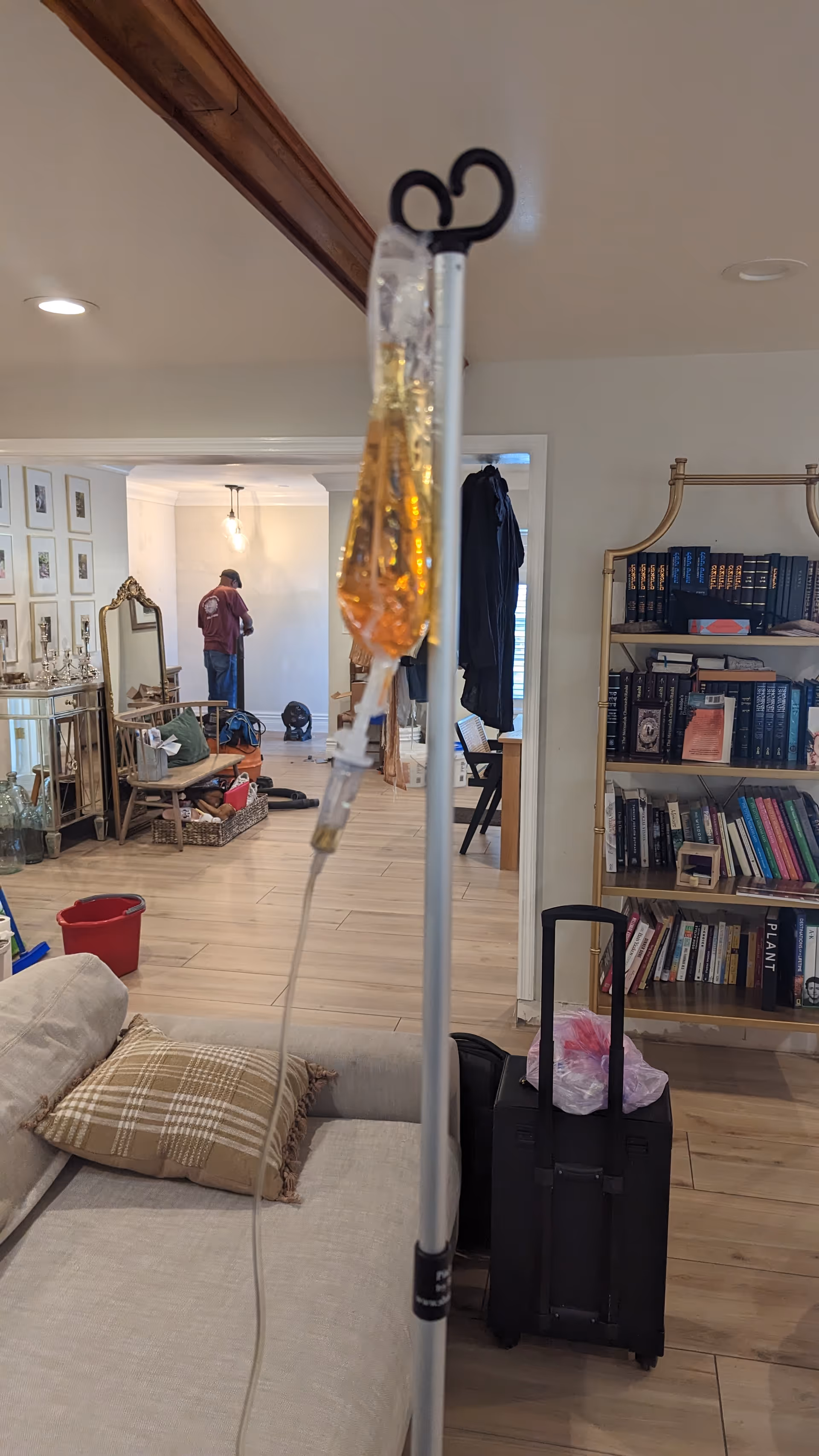 Indoor senior living lounge with IV stand, sofa, bookshelf, and a person in background.