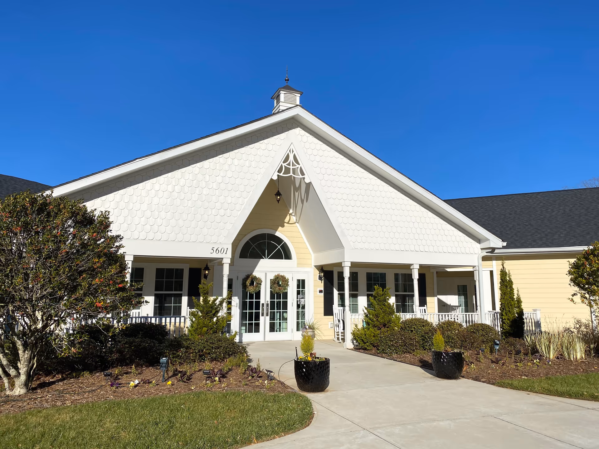 Front exterior view of a senior living facility building with a peaked roof, white siding, and a covered entrance. There are two wreaths on the glass double doors, shrubs and small trees on either side of the walkway, and a clear blue sky above.