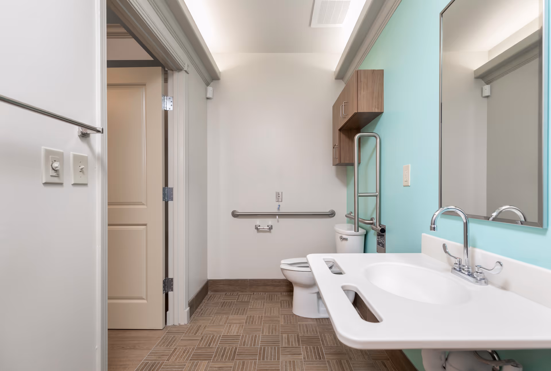 A clean and accessible bathroom featuring a white toilet with grab bars, a white sink with a wide countertop and a chrome faucet, a large mirror above the sink, and a wooden cabinet mounted on a light turquoise wall. The floor has a patterned tile design, and the walls are painted white and turquoise. The bathroom door is open, showing a beige door and door frame.