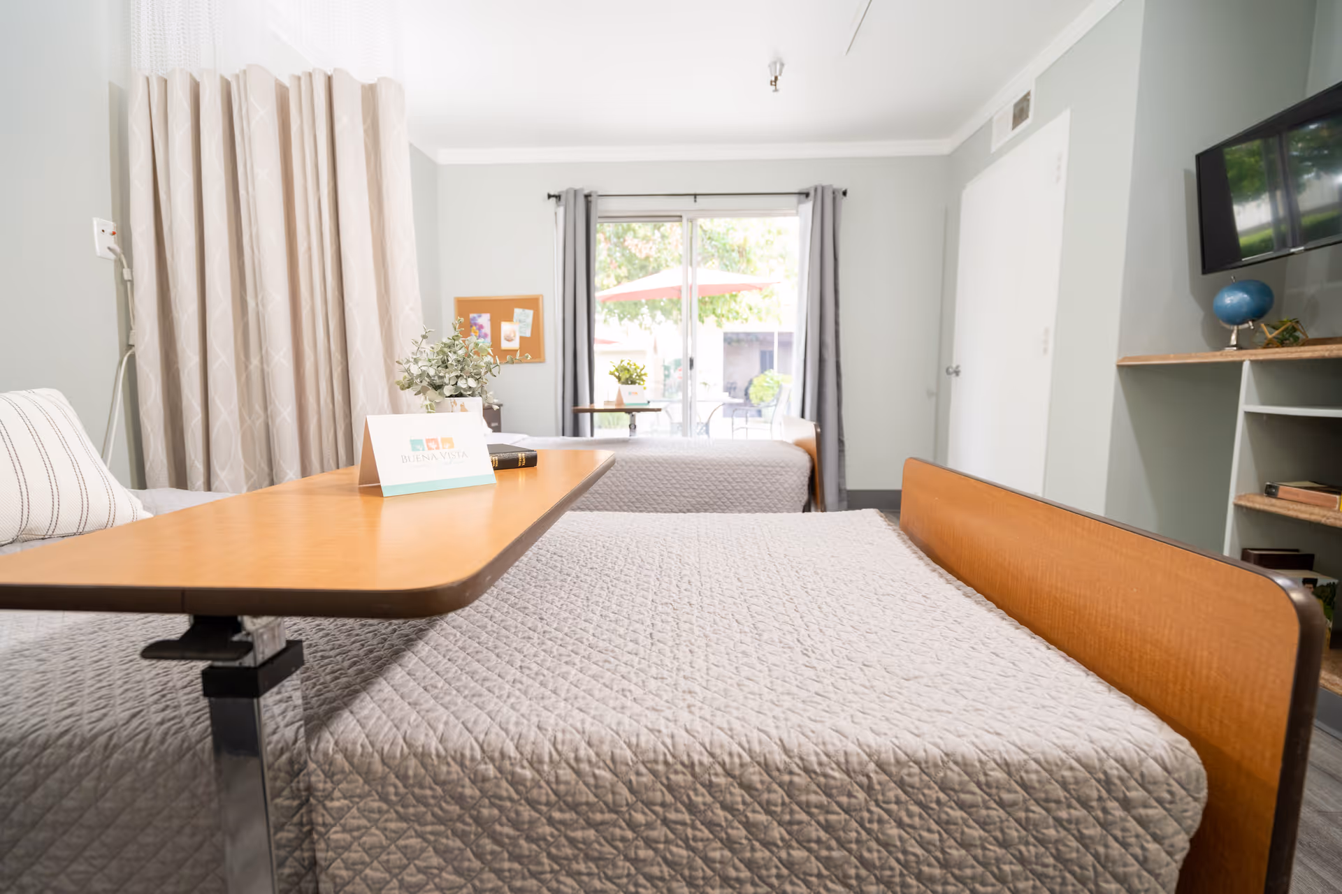 Two neatly made beds in a bright resident room with a desk and large window.