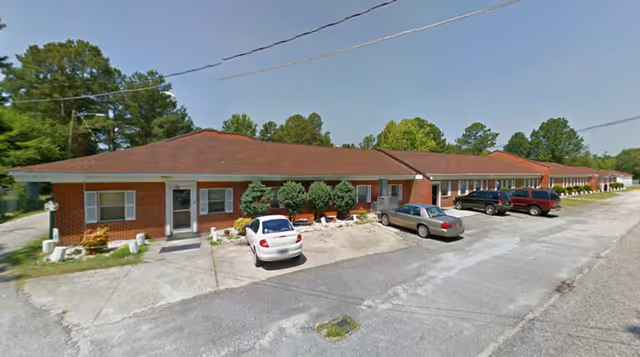 Exterior view of a single-story brick building with a red roof, several windows, and a parking area in front with multiple cars parked. There are small bushes and trees planted along the building.