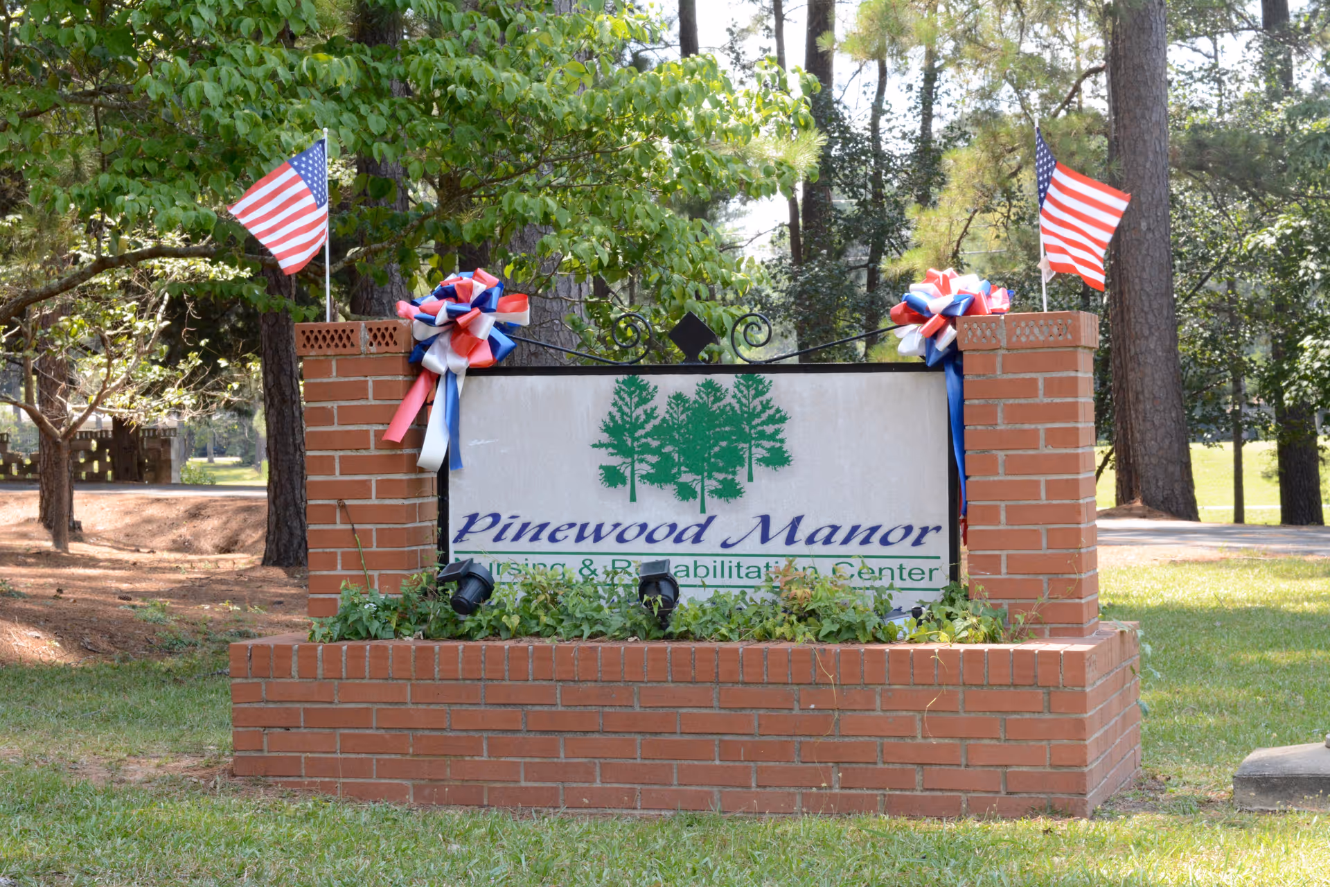 A brick sign for Pinewood Manor Nursing & Rehabilitation Center decorated with red, white, and blue bows and two small American flags, surrounded by greenery and trees in the background.