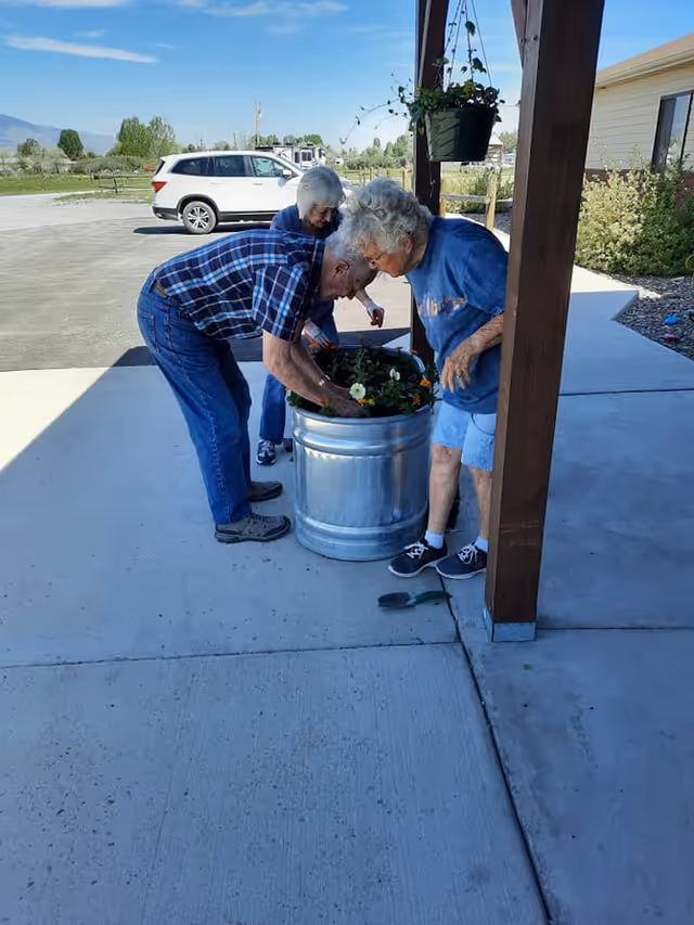 Three elderly individuals tending to a large metal planter filled with flowers on a concrete patio outside a building. A white SUV is parked nearby, and there is a hanging plant above the planter. The scene is set on a sunny day with clear skies.