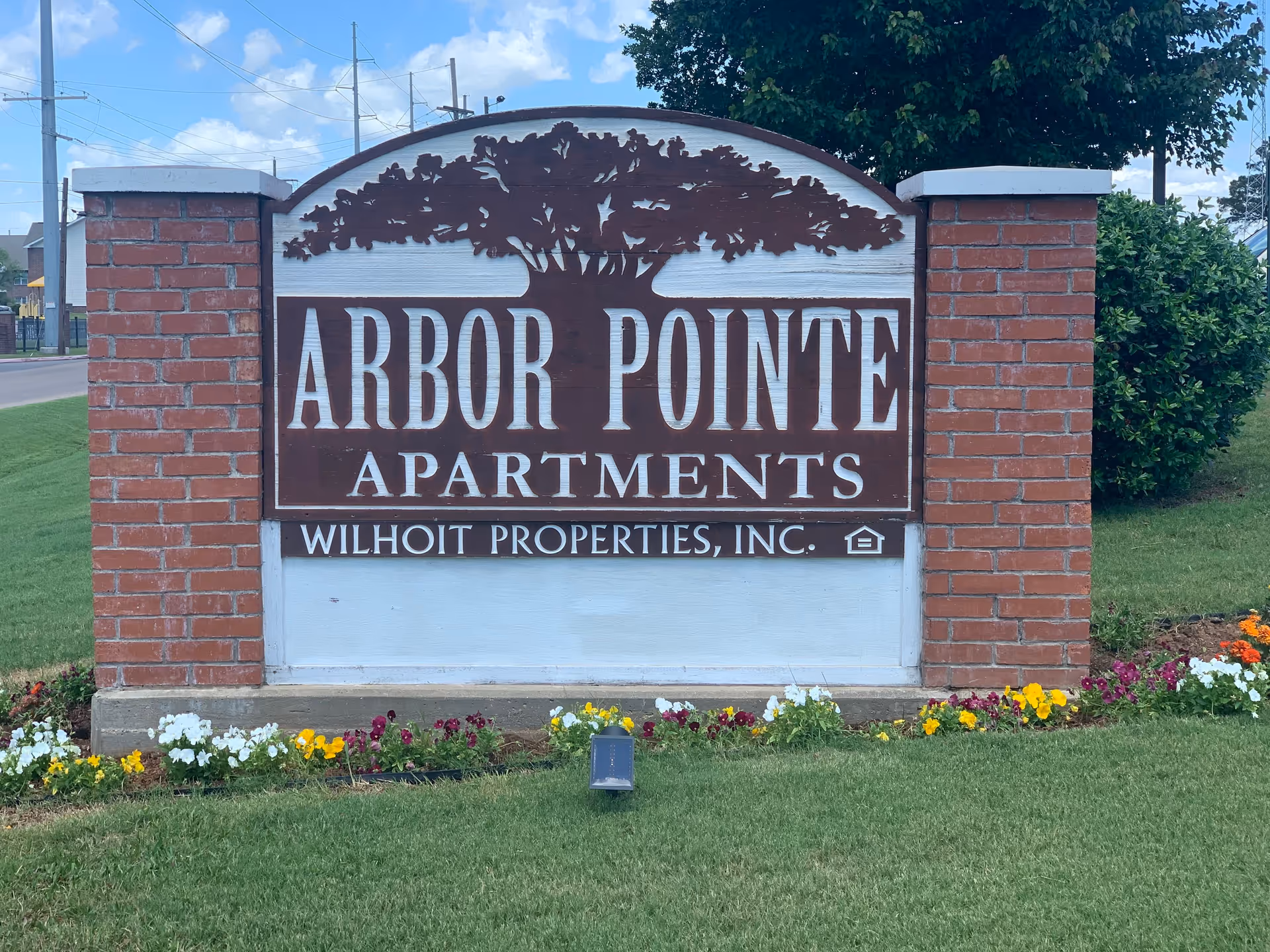 A large wooden sign for Arbor Pointe Apartments mounted between two brick pillars, with a tree graphic above the text. The sign is surrounded by a small flower bed with colorful flowers and green grass, under a partly cloudy blue sky.