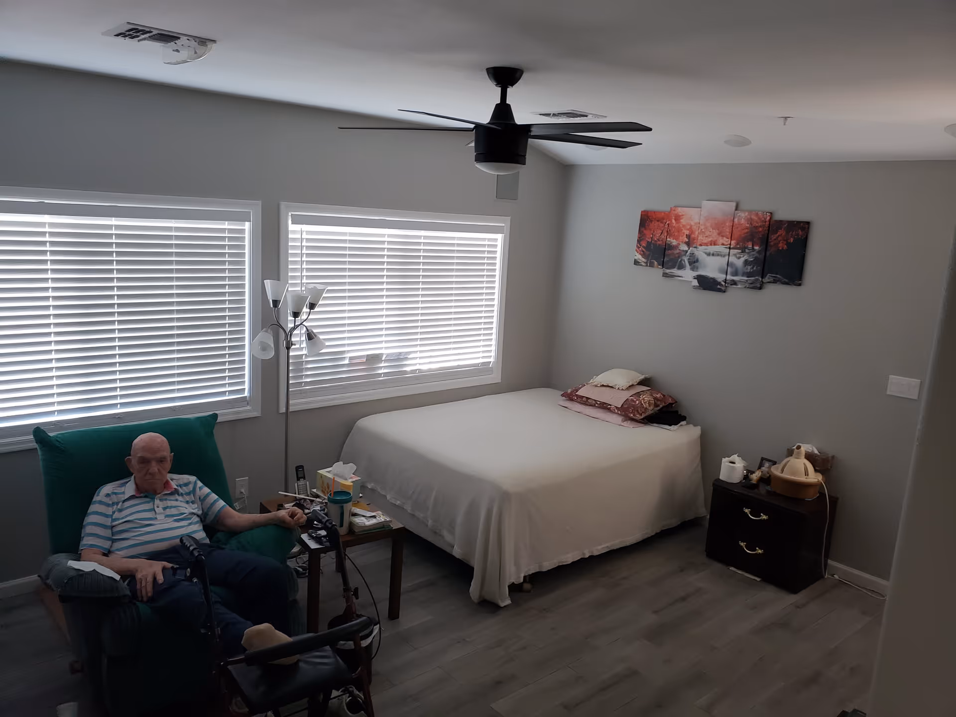 A simple bedroom with a bed, nightstand, windows with blinds and a man seated in a recliner under a ceiling fan.