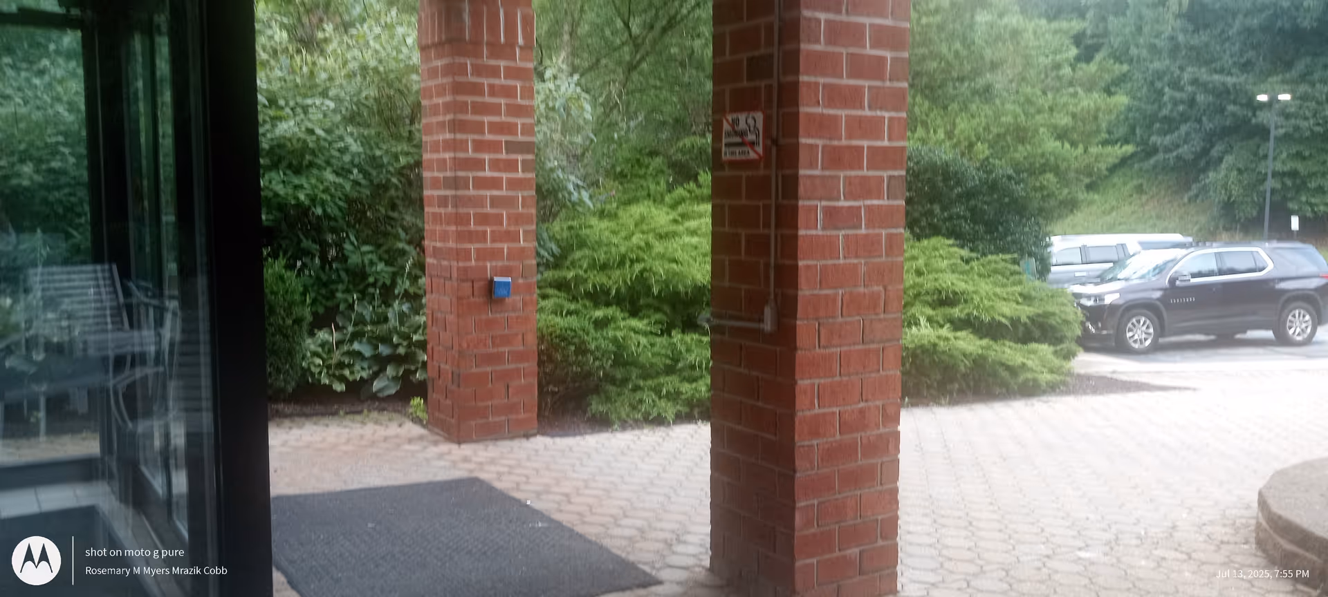Brick entrance columns frame glass doors with greenery and a parking lot.