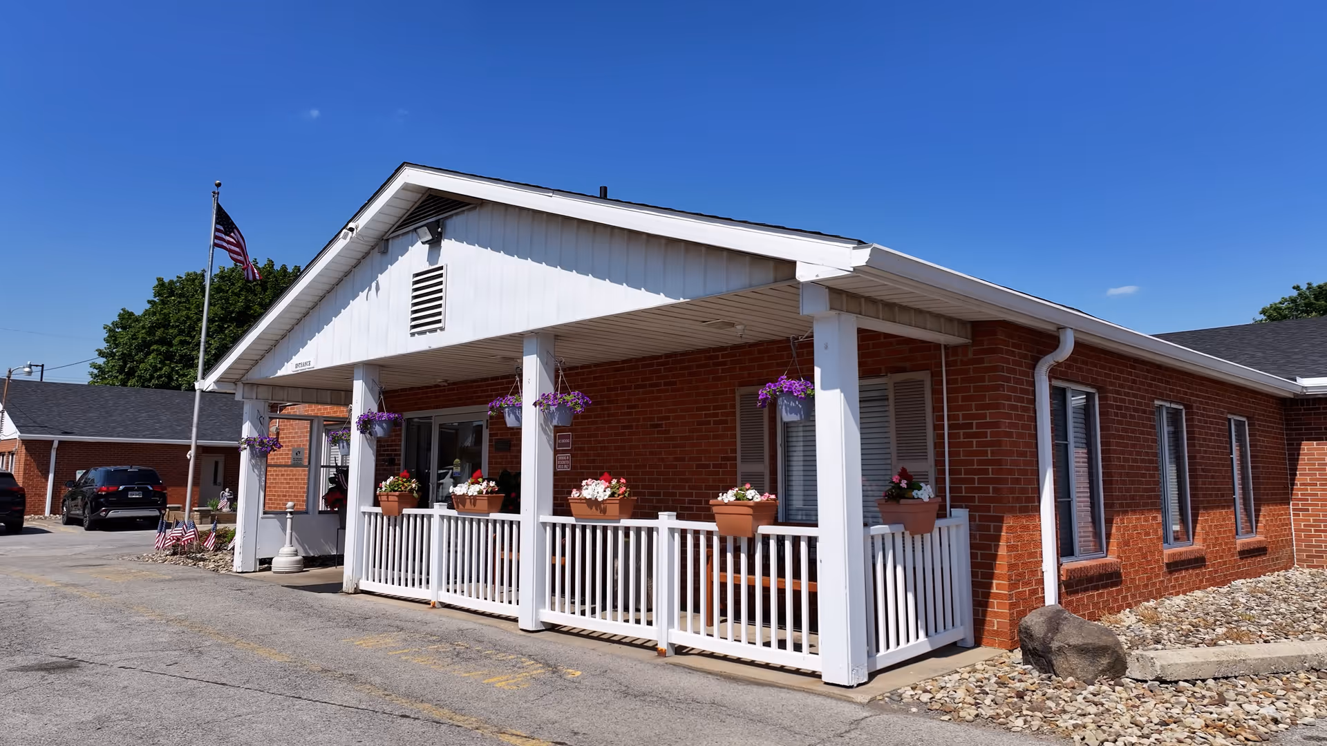 Front entrance of a single-story red brick building with a white covered porch, potted flowers, and an American flag.