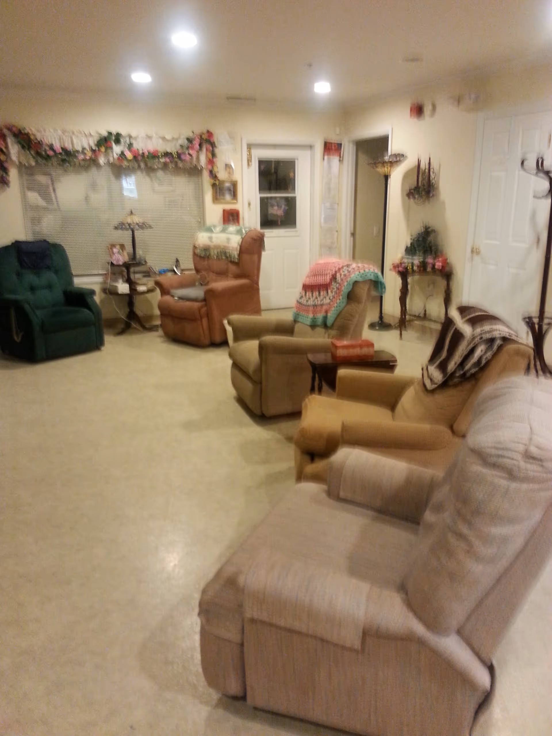 A cozy communal living room with several recliner chairs, side tables, and floral decorations near a door and window.