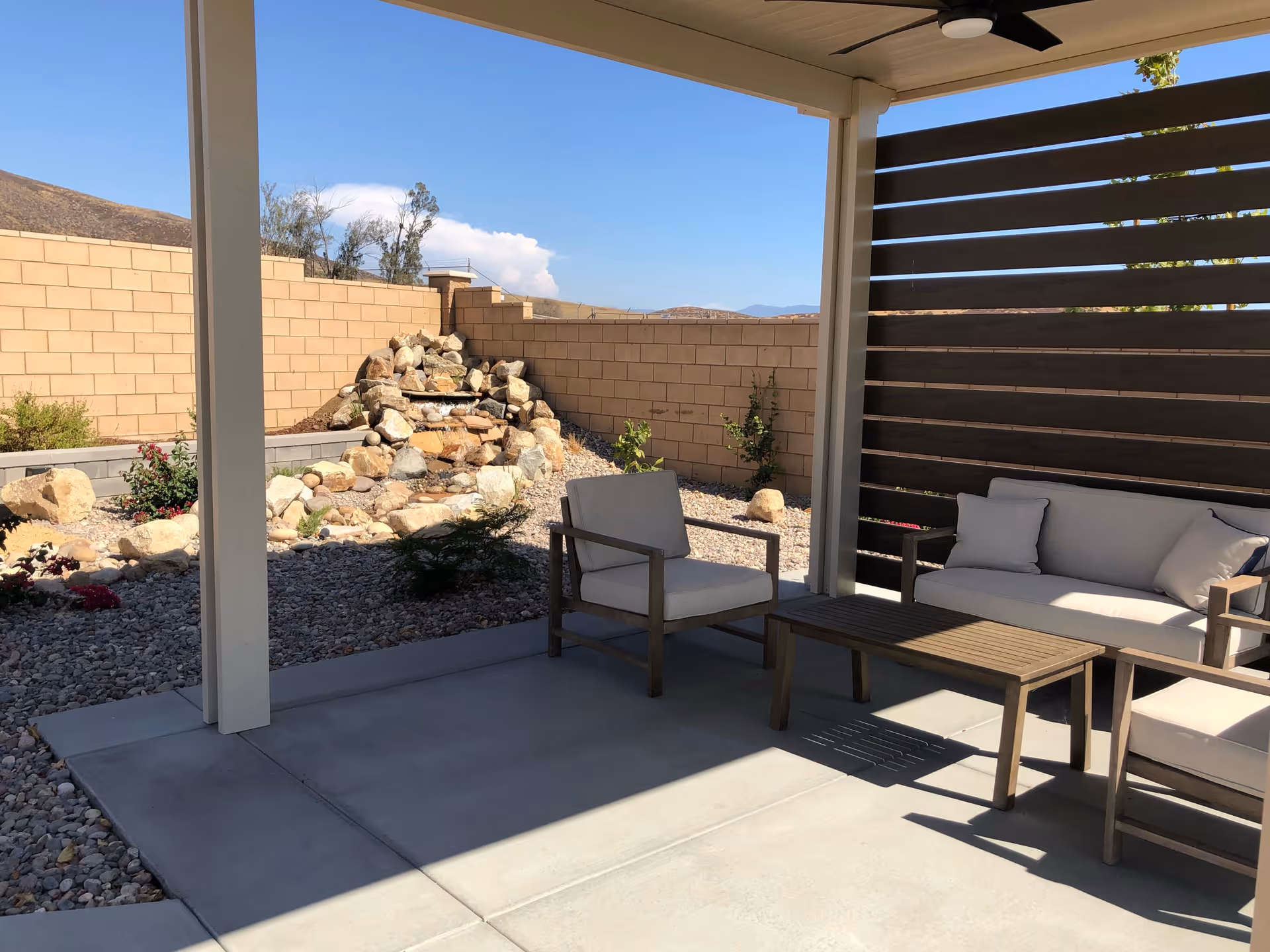 Outdoor patio area with cushioned seating including a sofa and two armchairs around a wooden coffee table. The patio is covered with a roof and has a privacy screen on one side. In the background, there is a rock waterfall feature and a stone wall enclosing the garden area with some plants and shrubs.