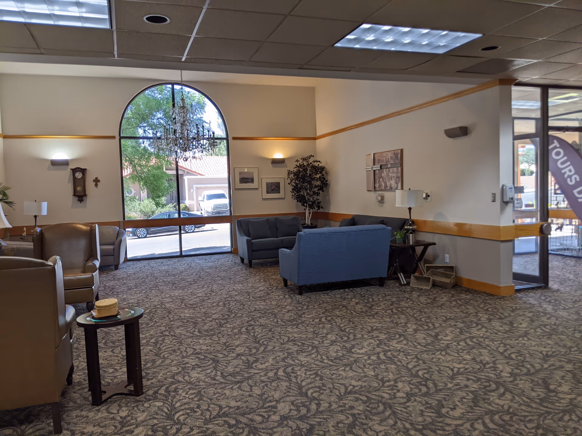Senior living facility lobby with blue sofas, arched window, chandelier, and patterned carpet.