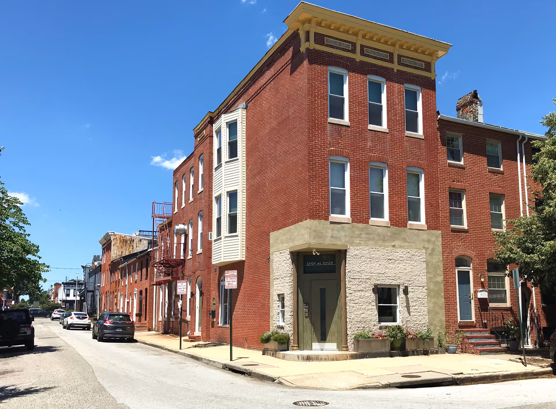 Three-story red-brick corner building with a greenish entrance on a sunny street lined with parked cars and a clear blue sky.