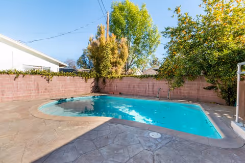 In-ground swimming pool in a fenced backyard with a stamped-concrete patio and surrounding trees.