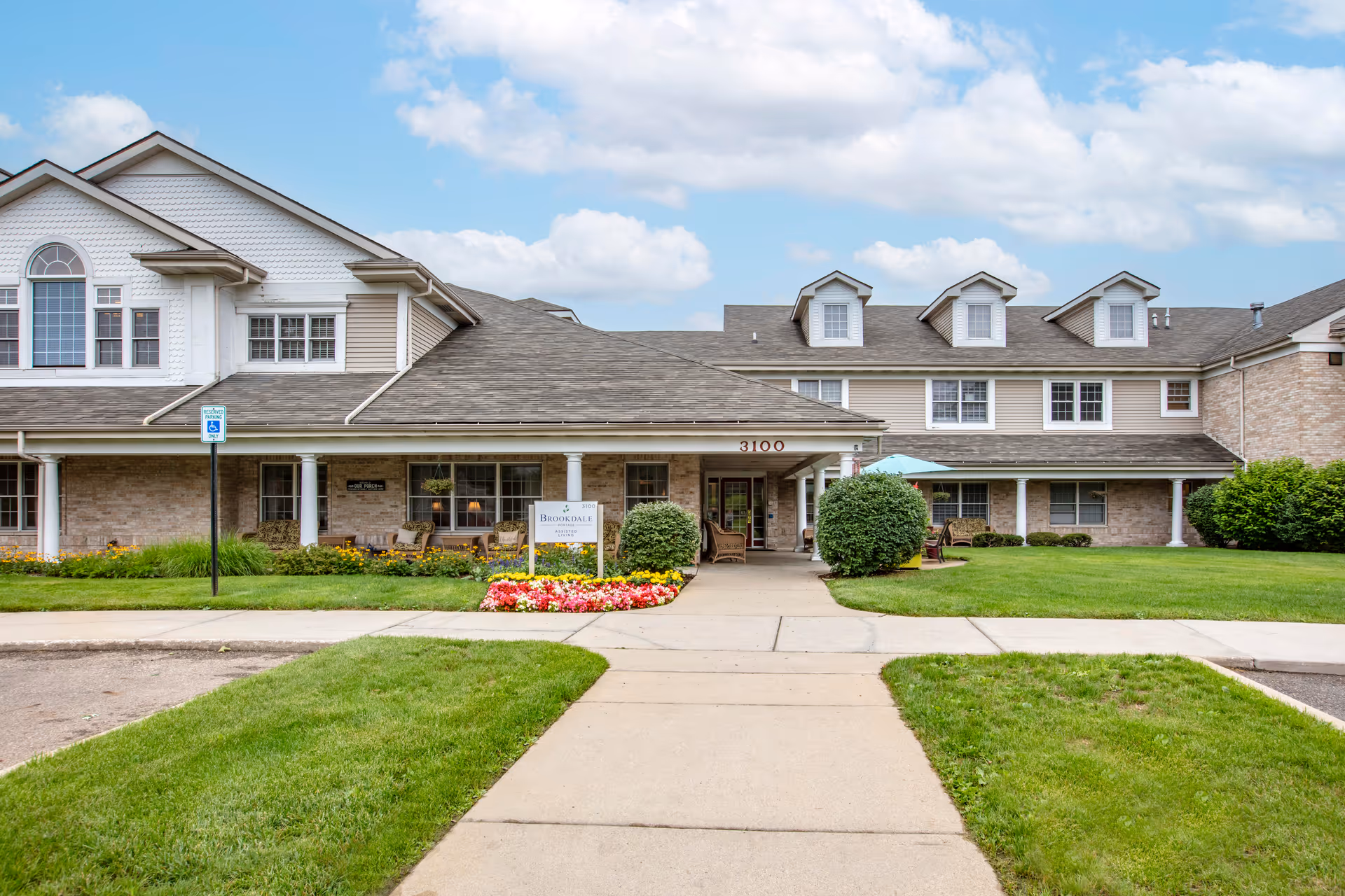 Front exterior view of Brookdale Portage Memory Care facility showing a two-story building with beige siding and brick accents, a covered entrance with seating, well-maintained green lawns, flower beds, and a clear blue sky with some clouds.