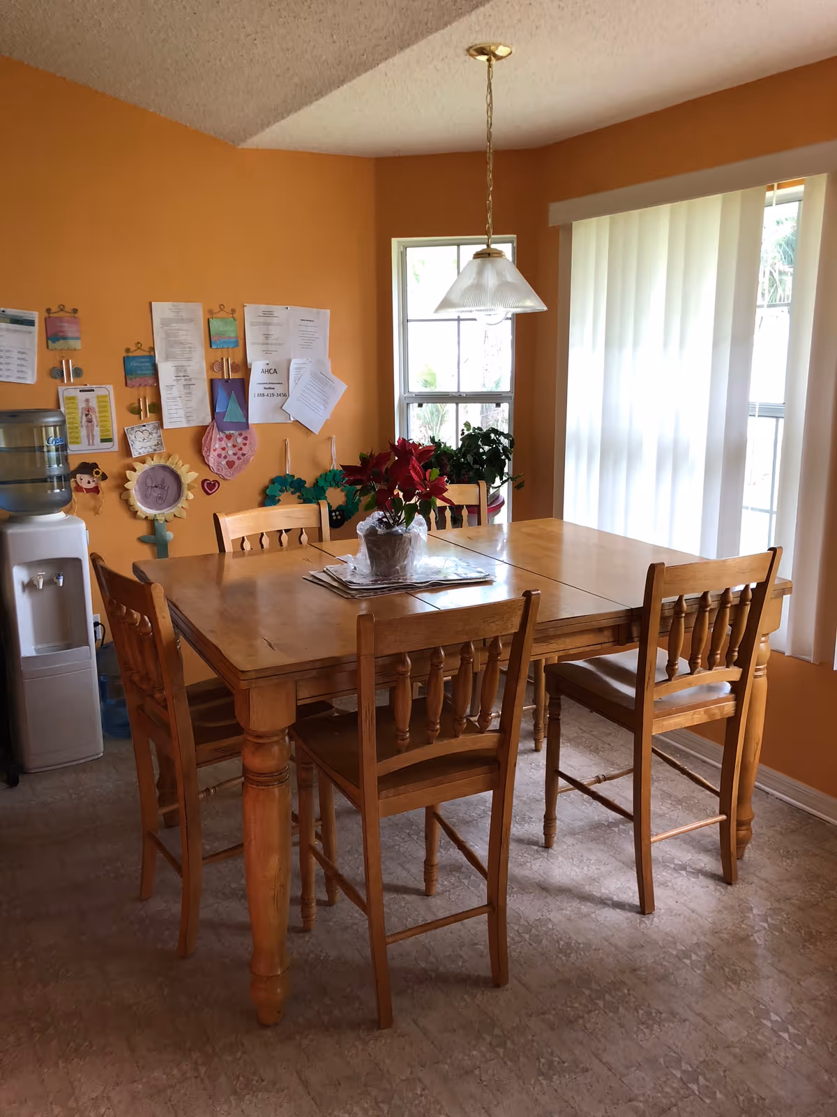 Orange-walled dining room with a wooden table, chairs, bulletin board, plants, and hanging light.