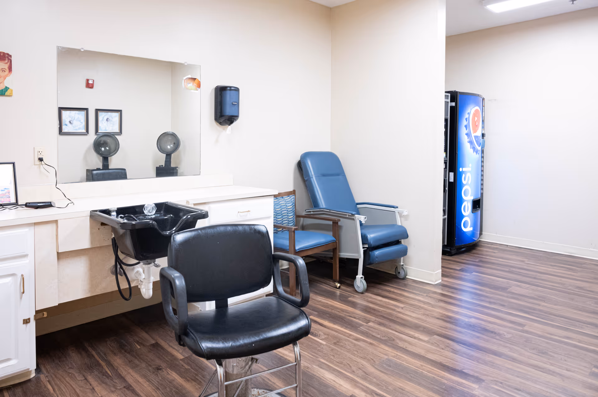Senior living facility hair salon area with sink, mirror, chairs, and Pepsi vending machine.