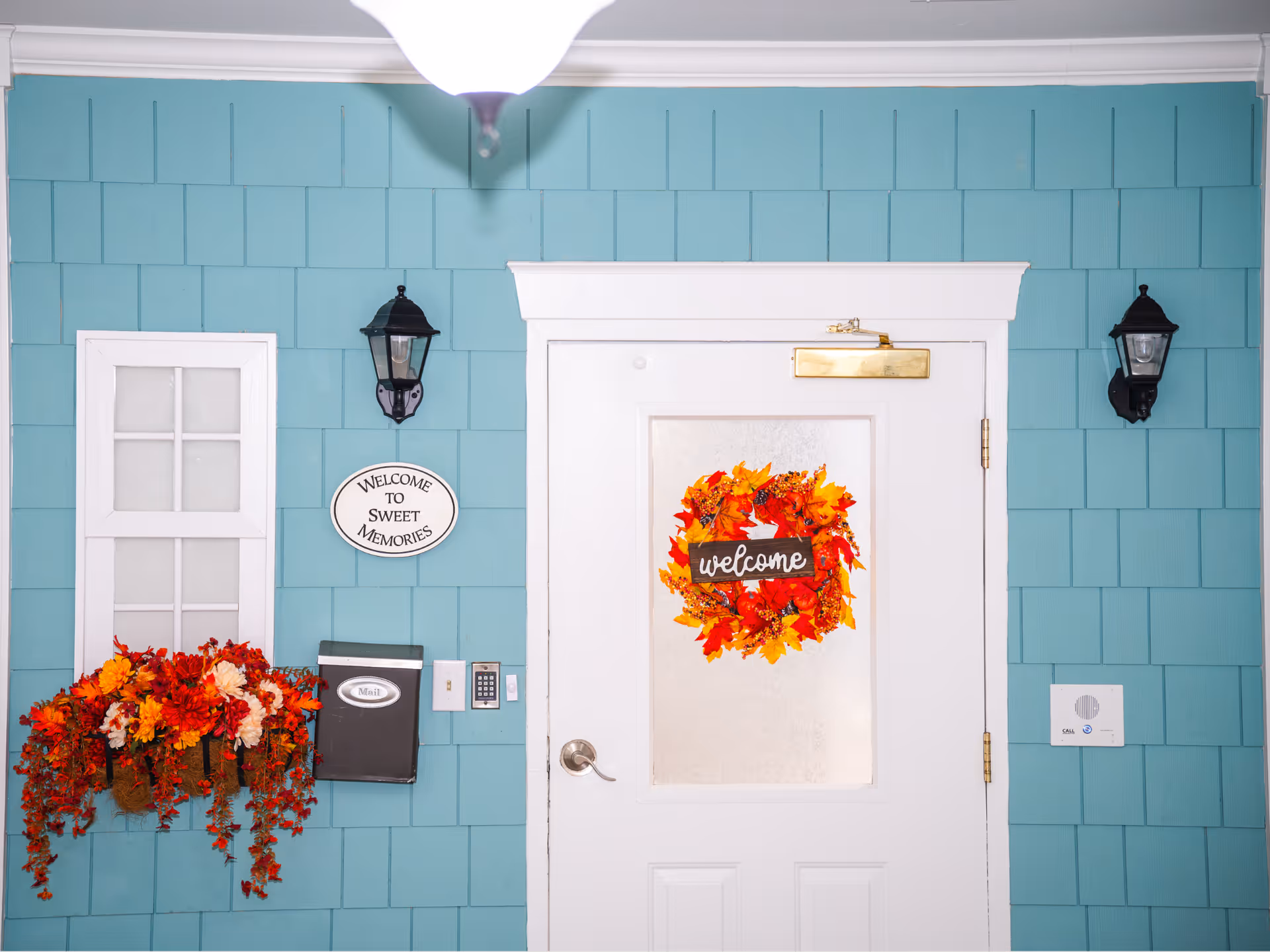 Blue exterior wall with white front door, lanterns, wreath, and autumn flowers.