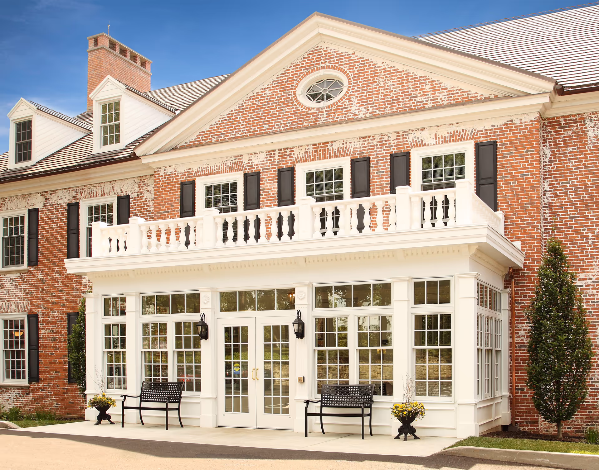 Front exterior of a red brick building with a white columned entrance, balcony, and benches by the doorway.