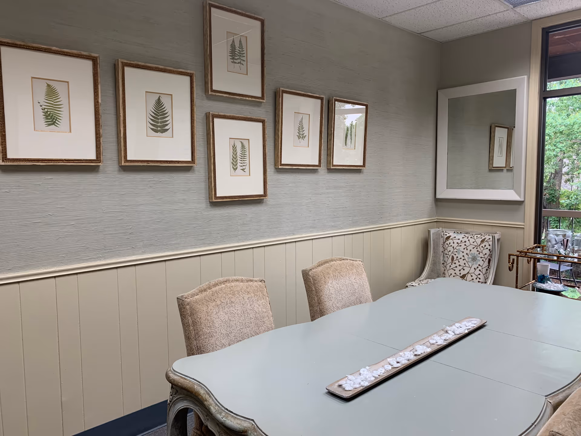 Indoor dining room with a pale blue table, framed fern art, and a mirror.