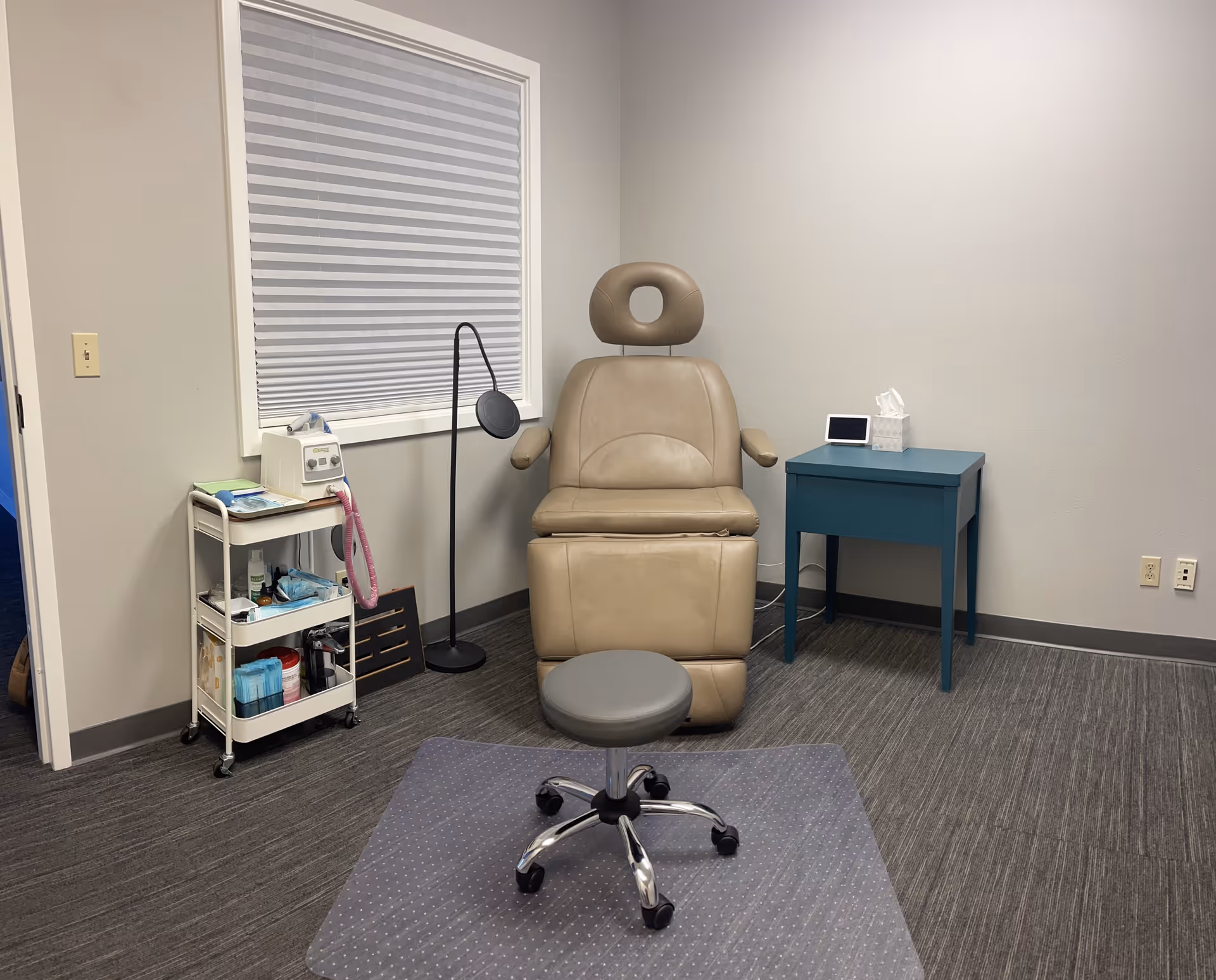Beige reclining treatment chair in a clinic room with a medical cart and blue table.