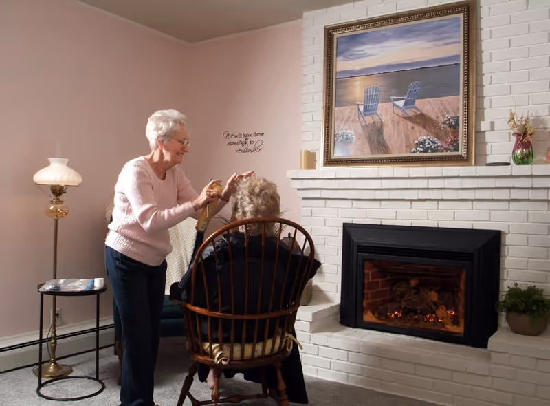 An elderly woman standing and styling the hair of another elderly woman seated in a wooden chair in a cozy living room with a white brick fireplace, a painting of two chairs on a dock above the fireplace, a floor lamp, and a small side table.