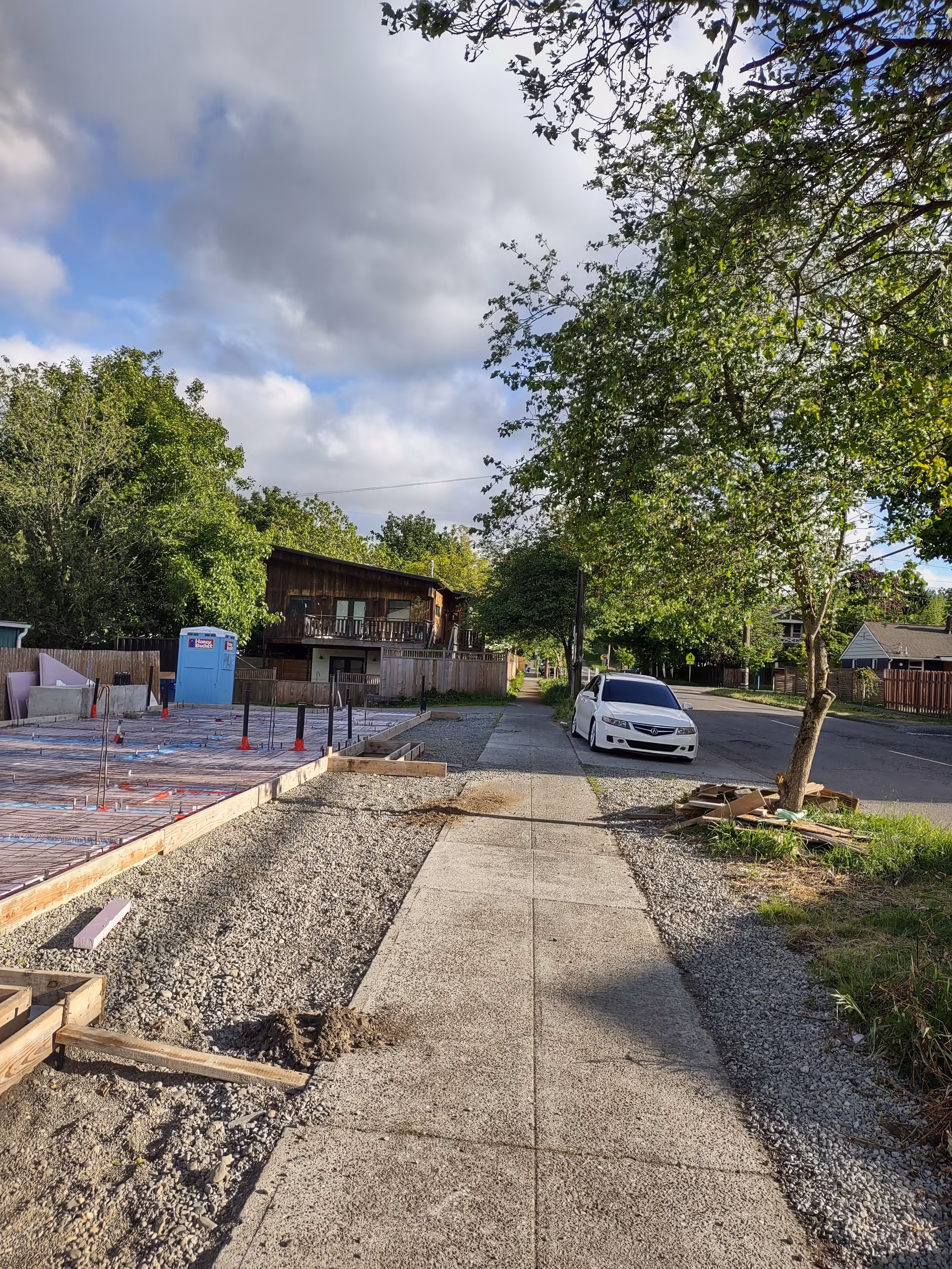 Residential street with ongoing construction on the left, a white car on the road, trees lining.