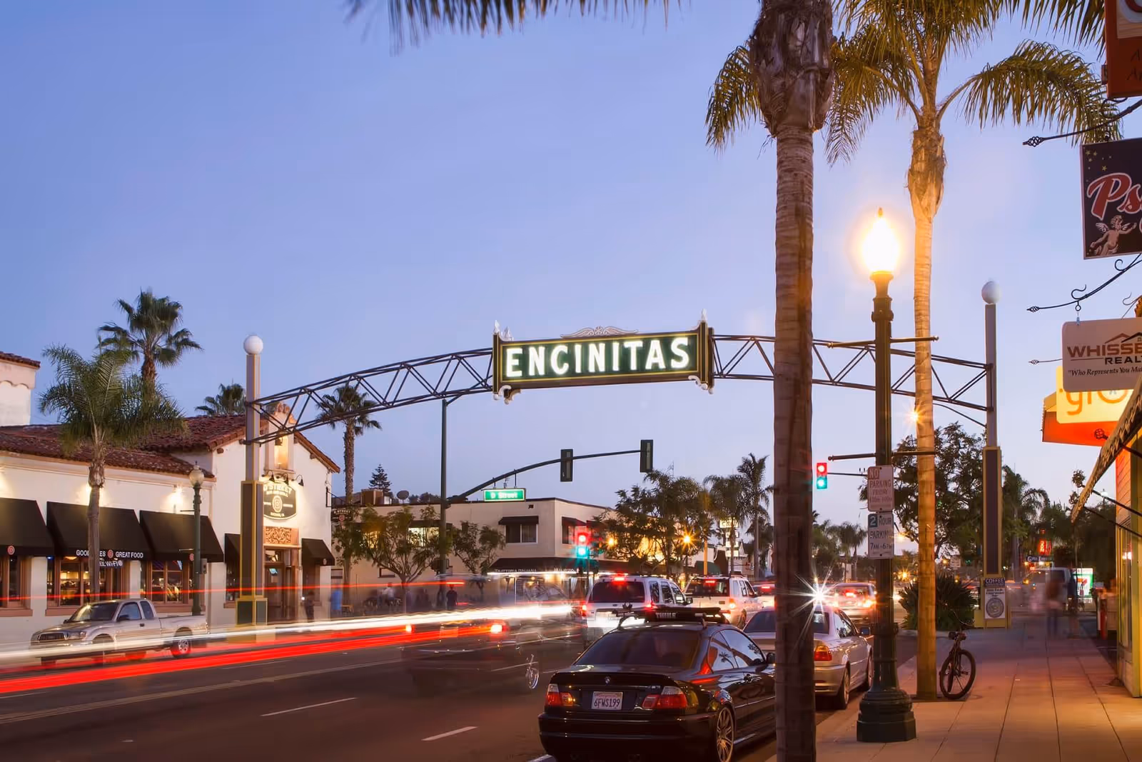 Urban street scene with palm trees, cars, and an Encinitas arch sign at dusk.